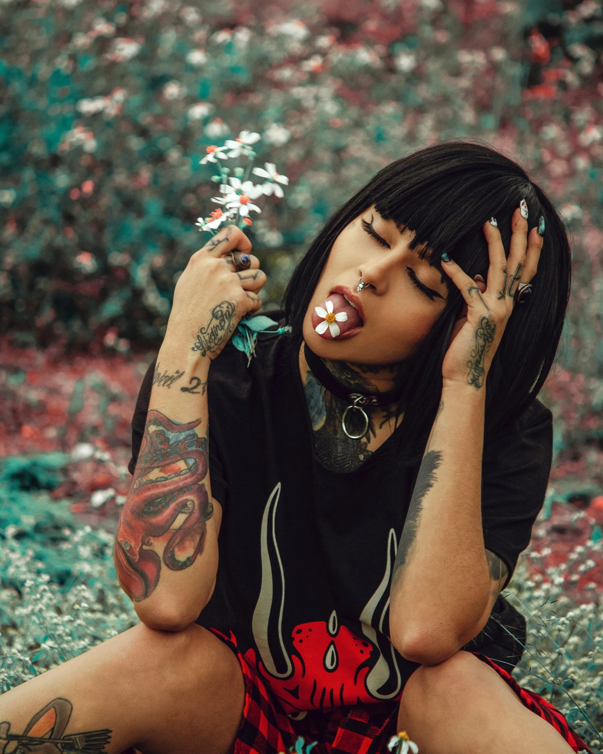 Goth woman with dark bob haircut and multiple tattoos, sitting in a field and holding a flower in her mouth.