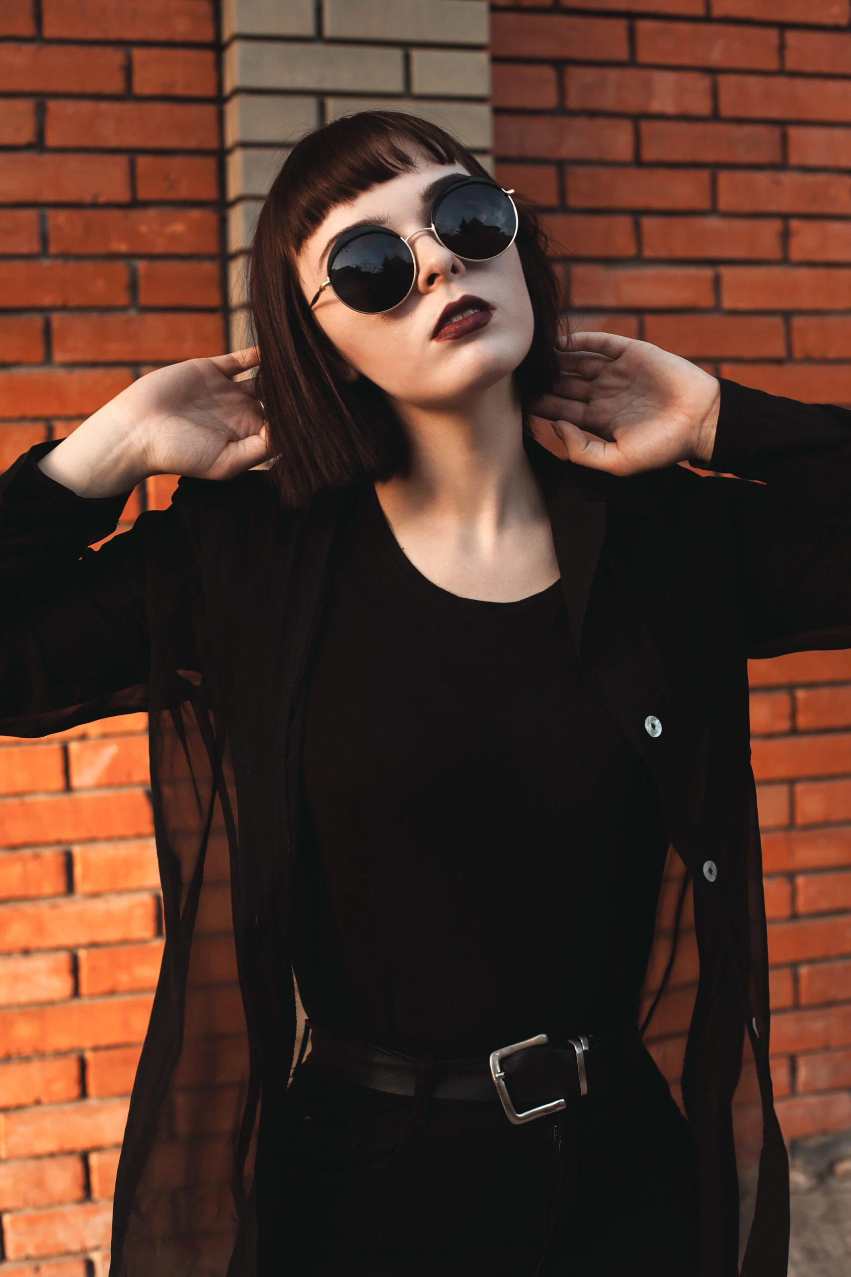 Stylish woman with bob haircut and round sunglasses standing in front of a brick wall, showcasing the trend of wearing sunglasses at night.