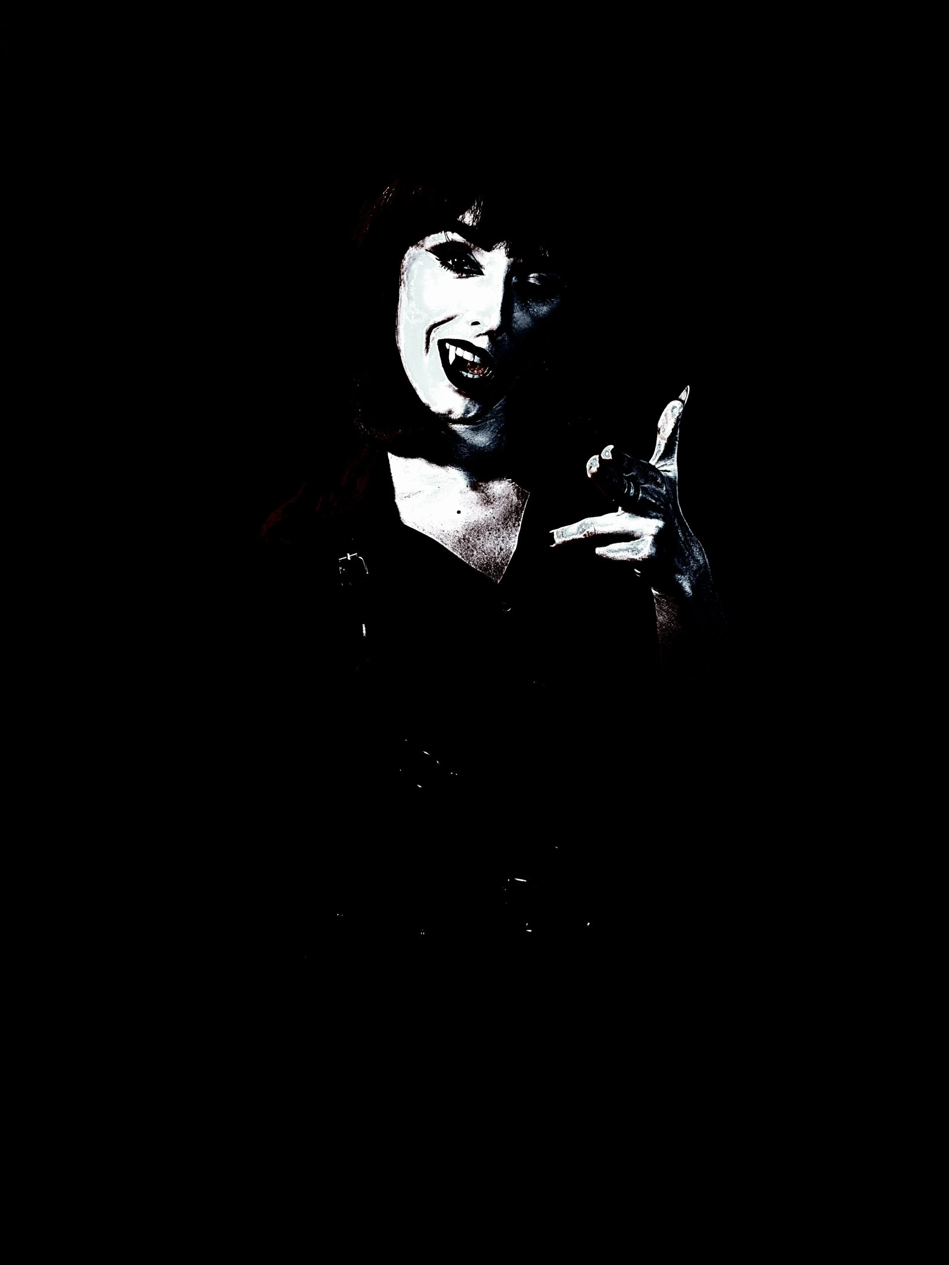 Gothic woman with vampire fangs, striking a playful pose in low light.