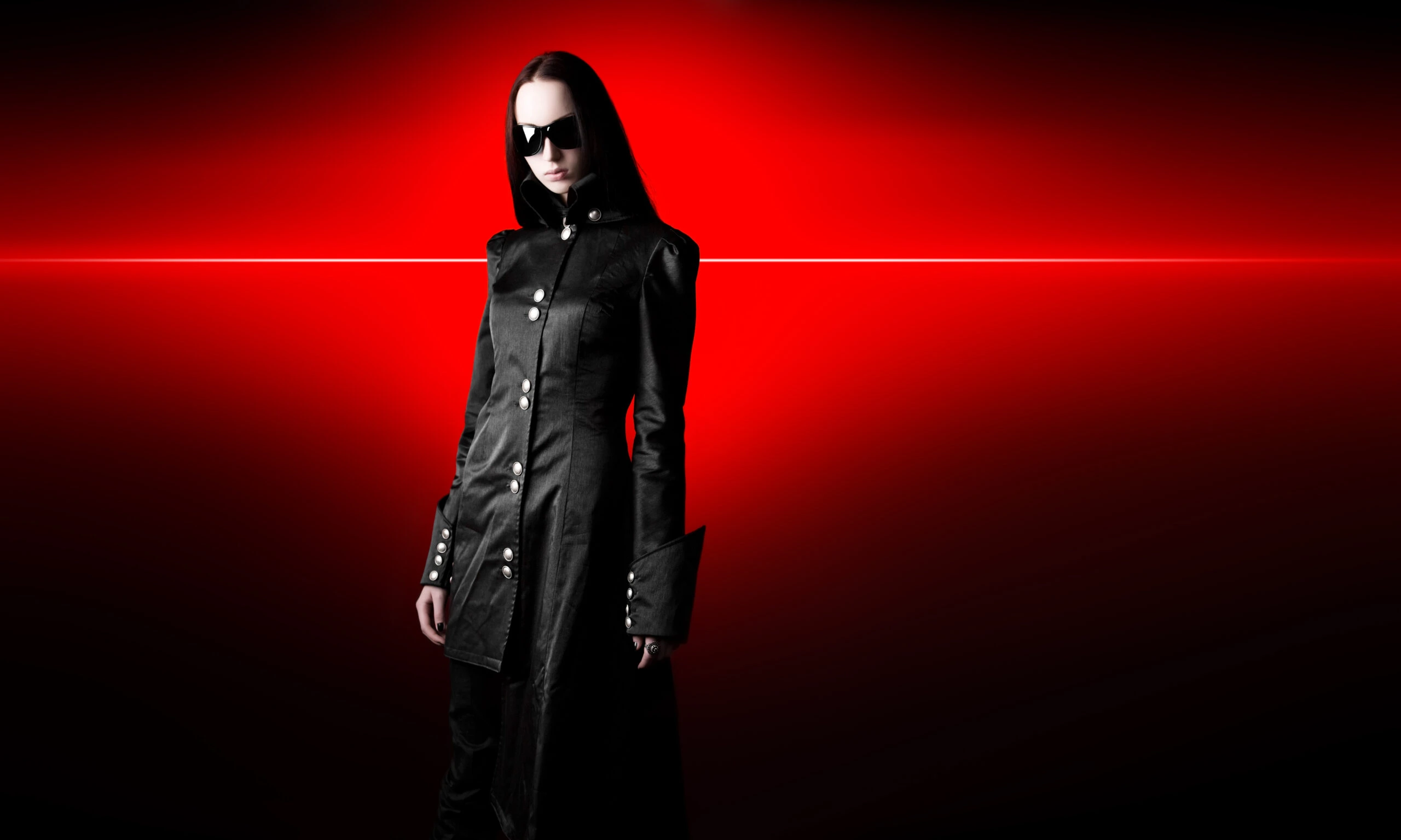 Gothic woman in a long coat and sunglasses, standing against a red background, illustrating the mystique of wearing sunglasses at night.