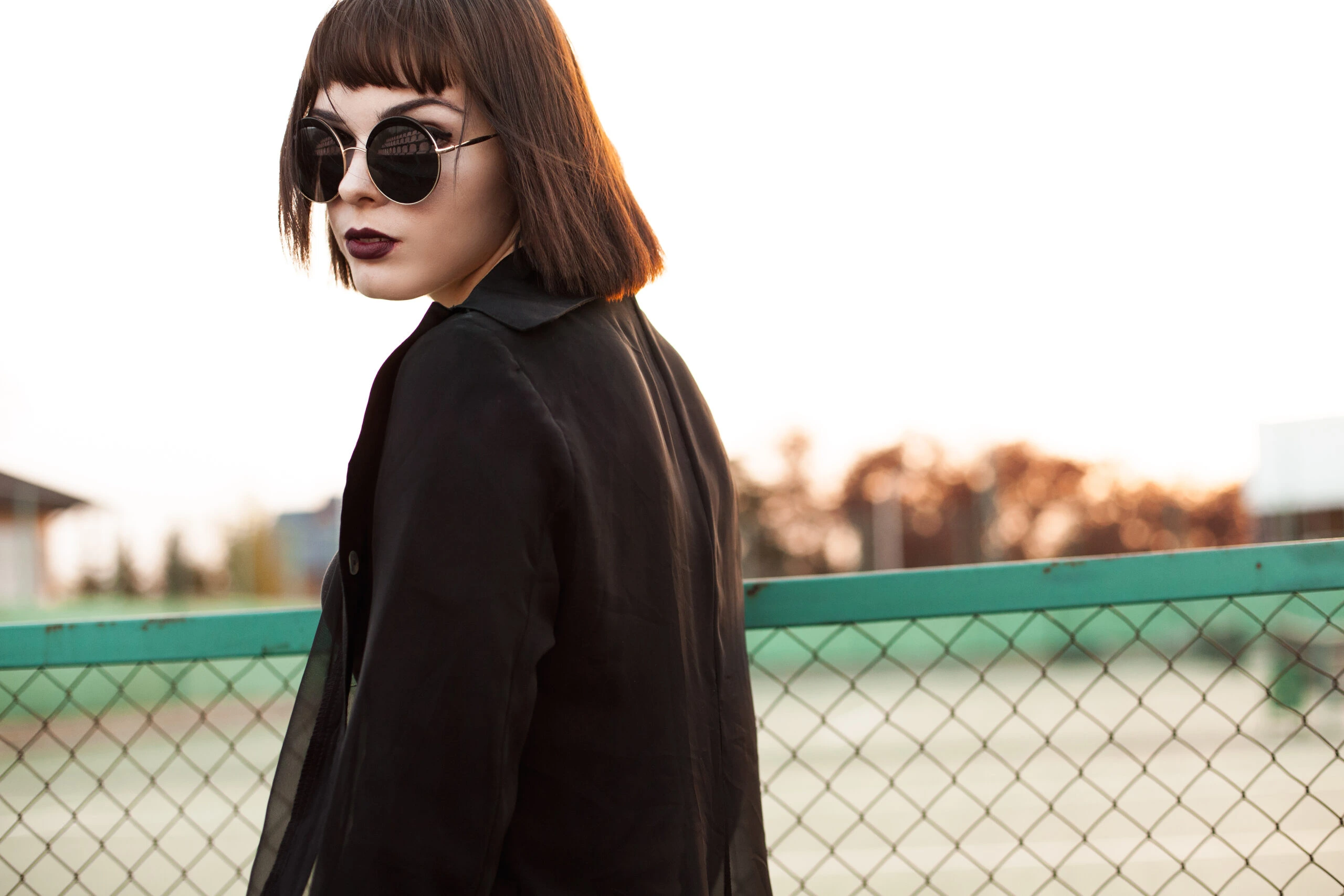 Woman in black coat and sunglasses, standing by a fence, highlighting the fashion-forward trend of wearing sunglasses at night.
