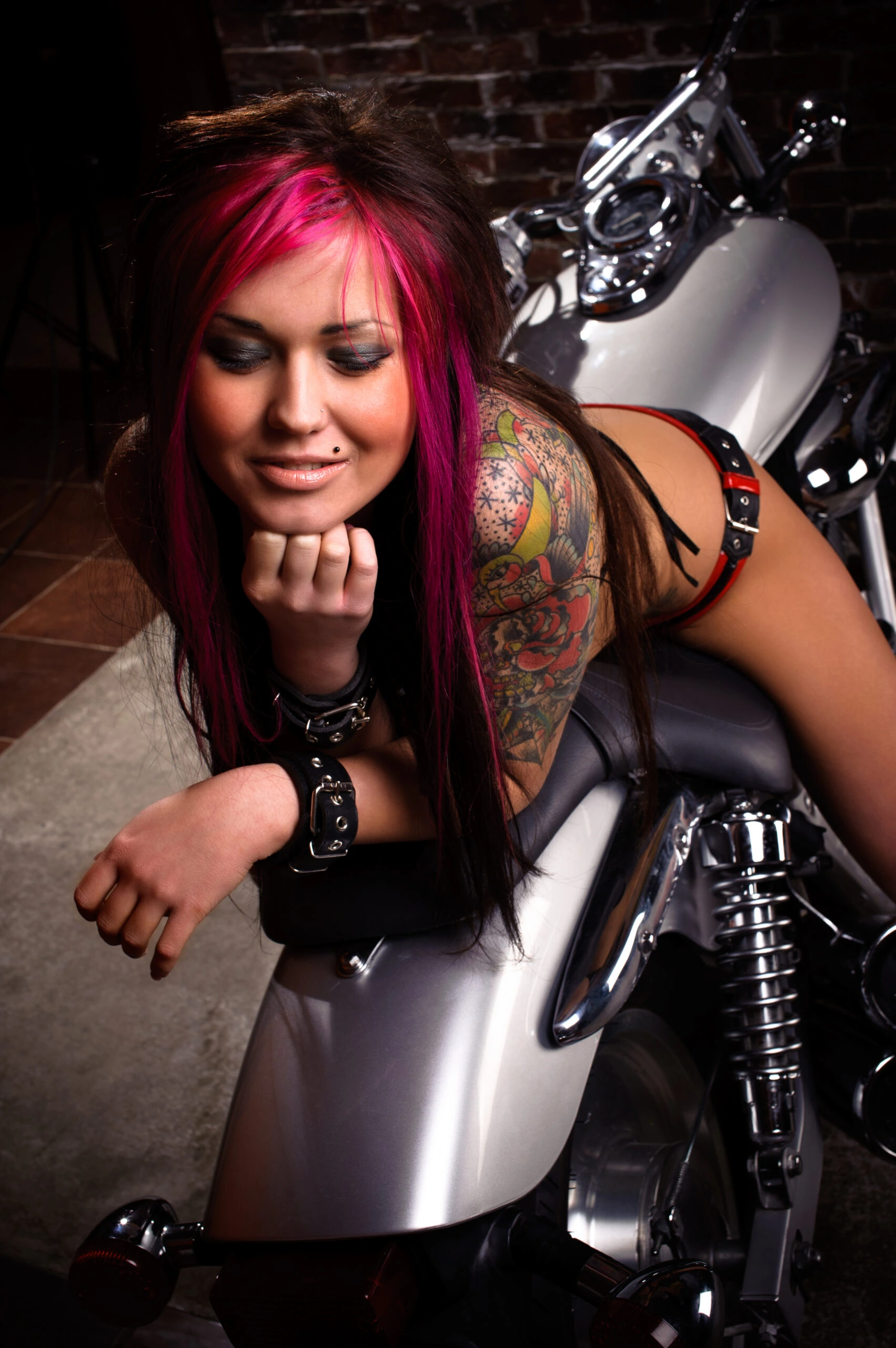 Goth woman with pink highlights and intricate tattoo, posing on a motorcycle in a dark, industrial setting.