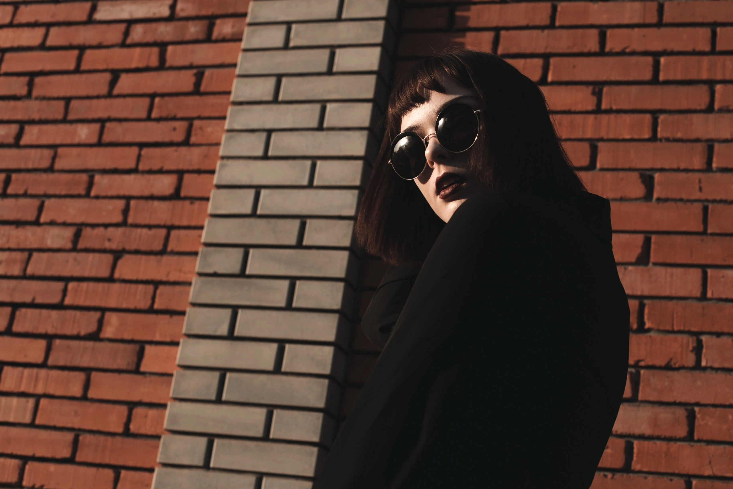 Woman in a black outfit and round sunglasses posing against a brick wall, illustrating the style of wearing sunglasses at night.