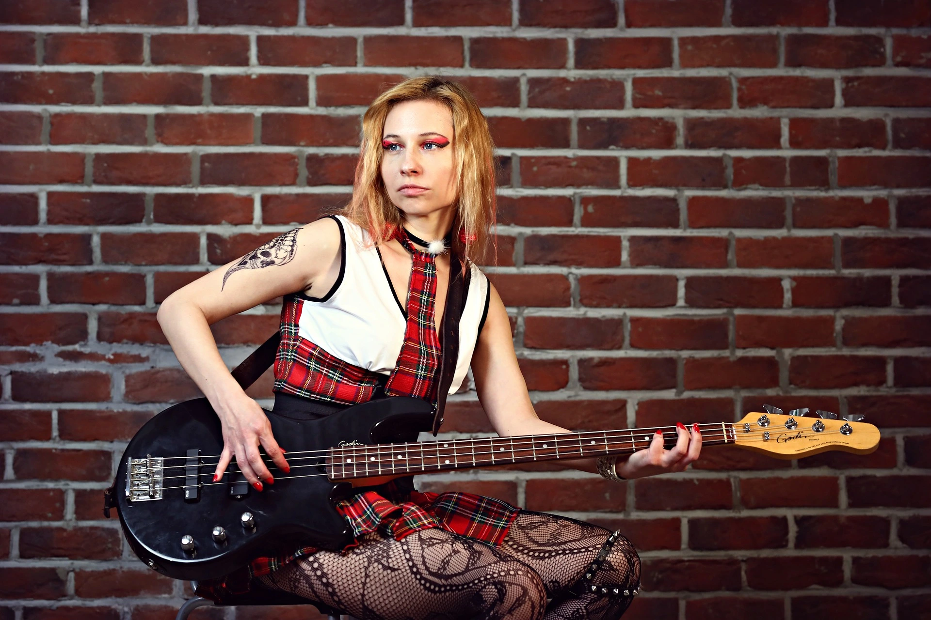 Woman with a punk-inspired look, playing a bass guitar in front of a brick wall, showcasing a gothic plaid outfit and tattoos.