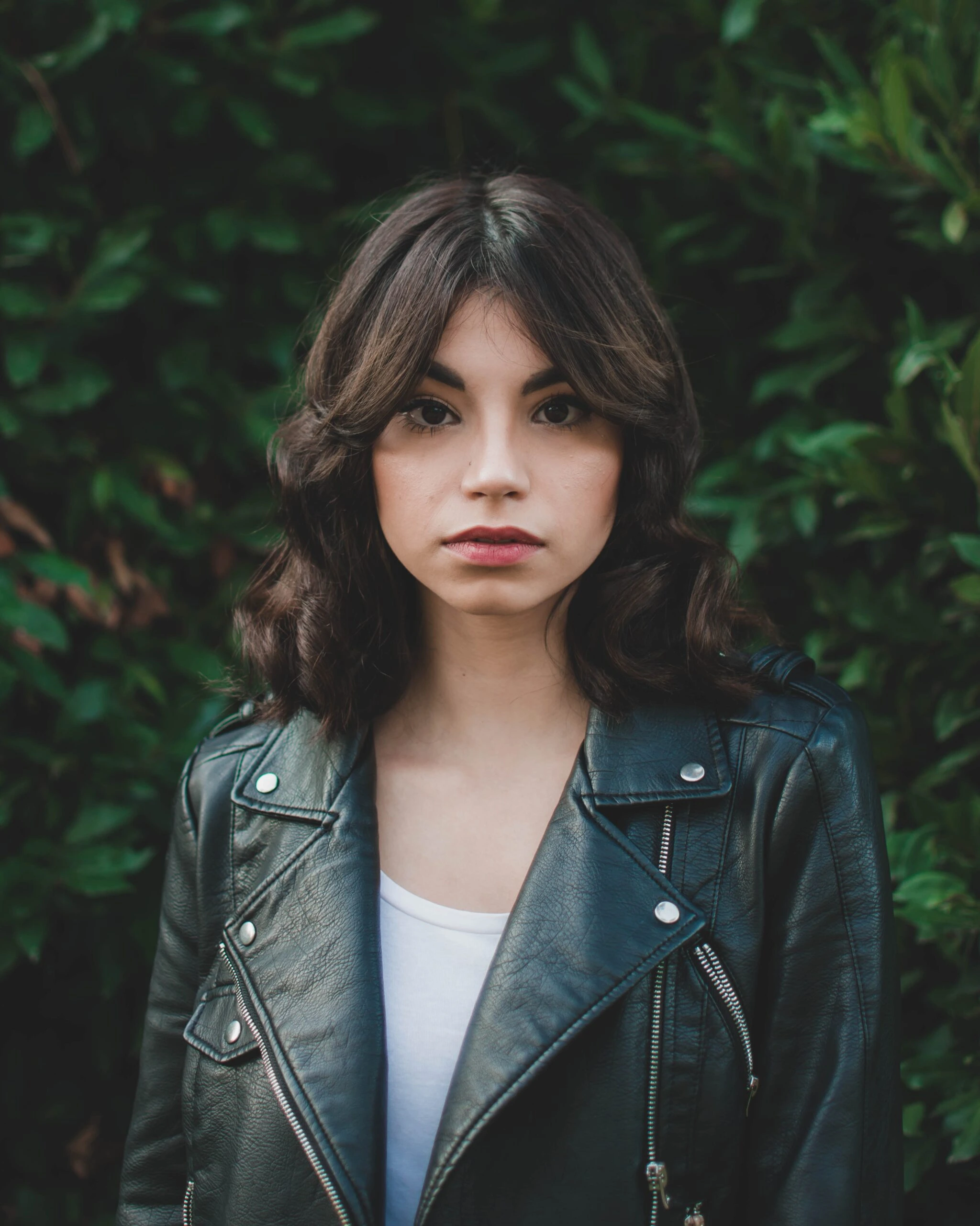 Portrait of a young woman with dark hair and a leather jacket standing against a leafy green background.