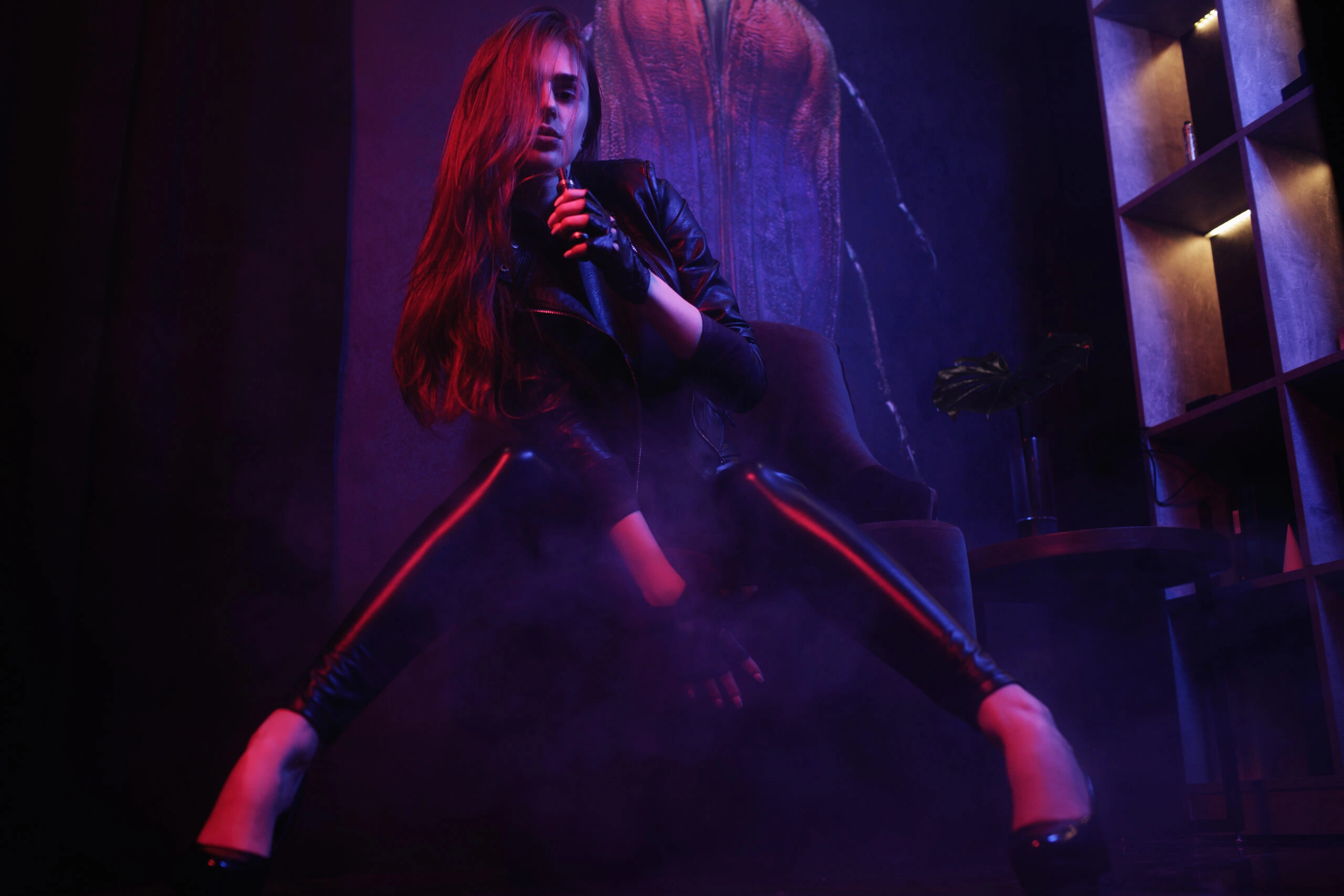 Gothic woman in a leather jacket and leggings, striking a confident pose in a dark, moody environment.