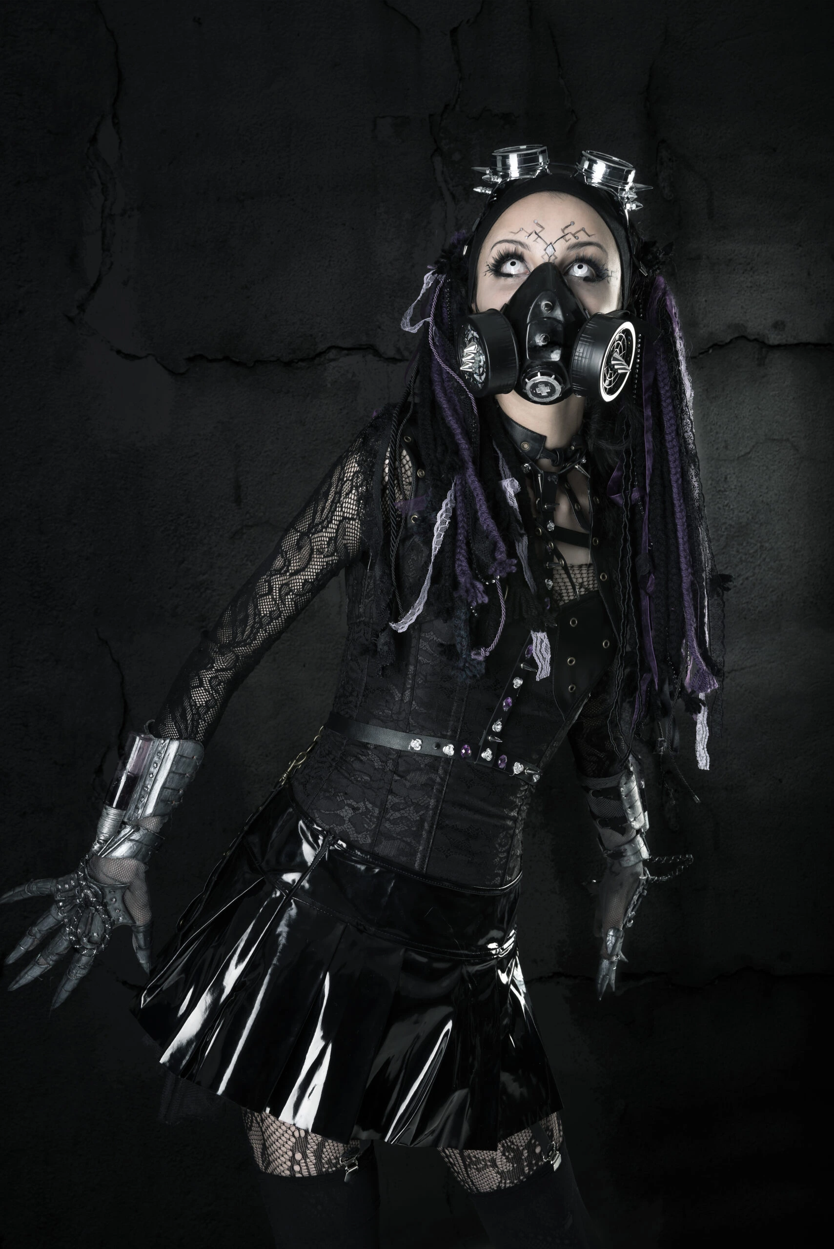 Cyber goth woman in a black lace over bust corset, gas mask, and metallic accessories against a cracked wall background.