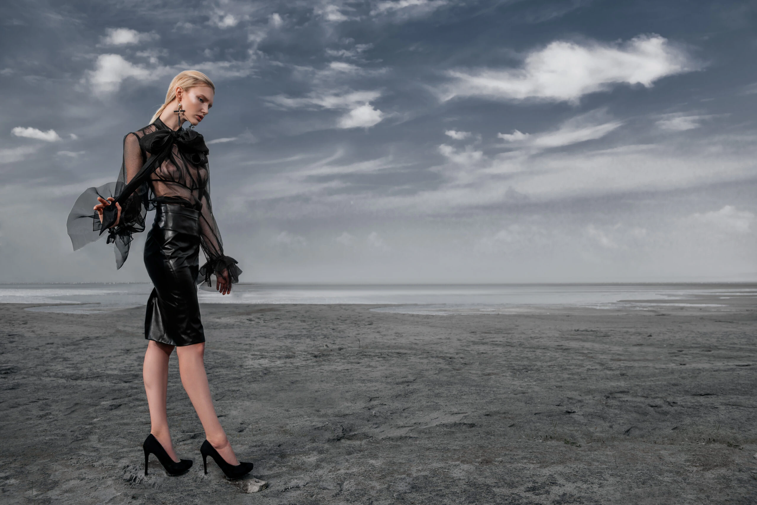 Woman in a sheer Gothic blouse and leather skirt, standing on a desolate beach.