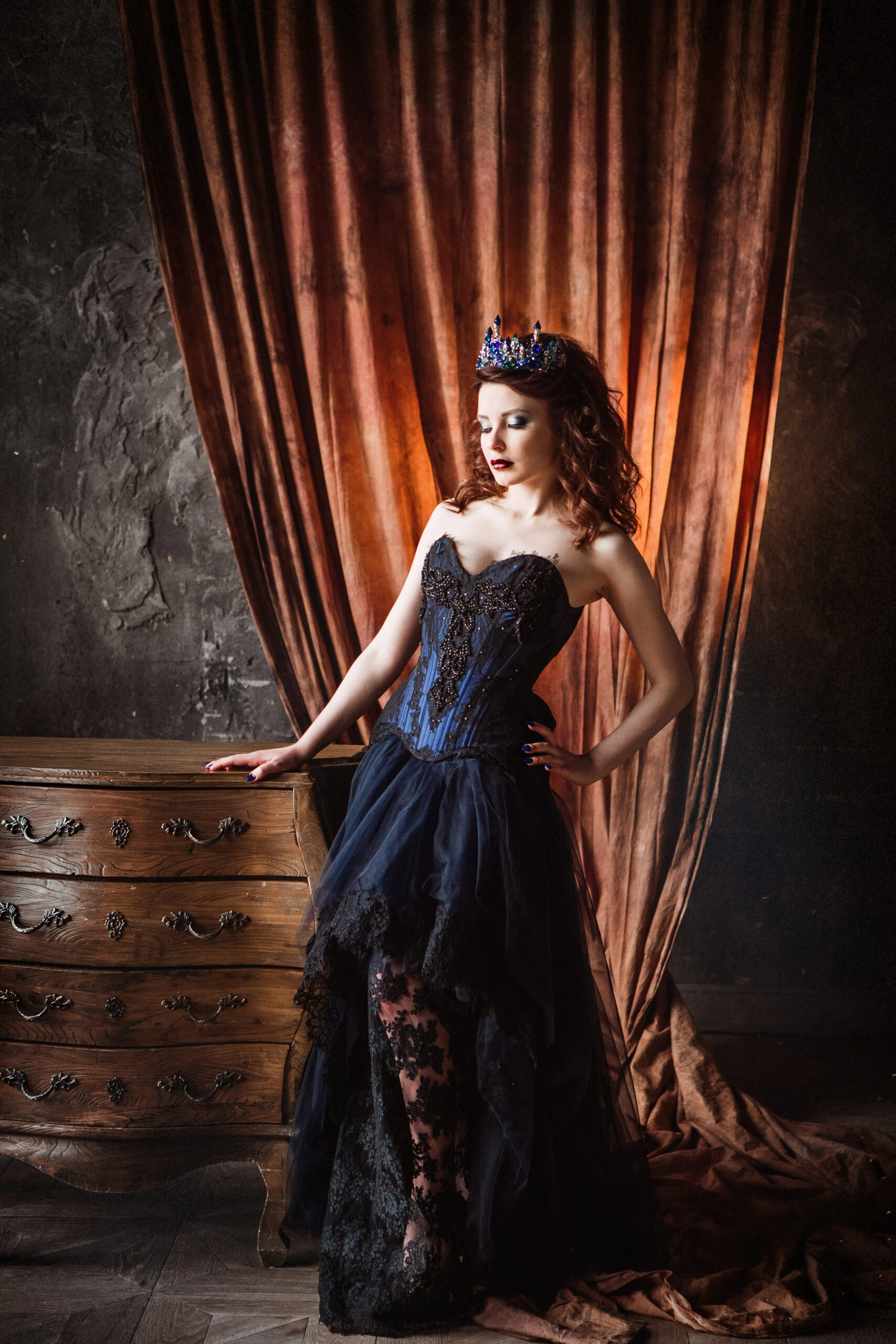 Woman in an elegant blue over bust corset gown standing beside a vintage wooden dresser with dramatic lighting.