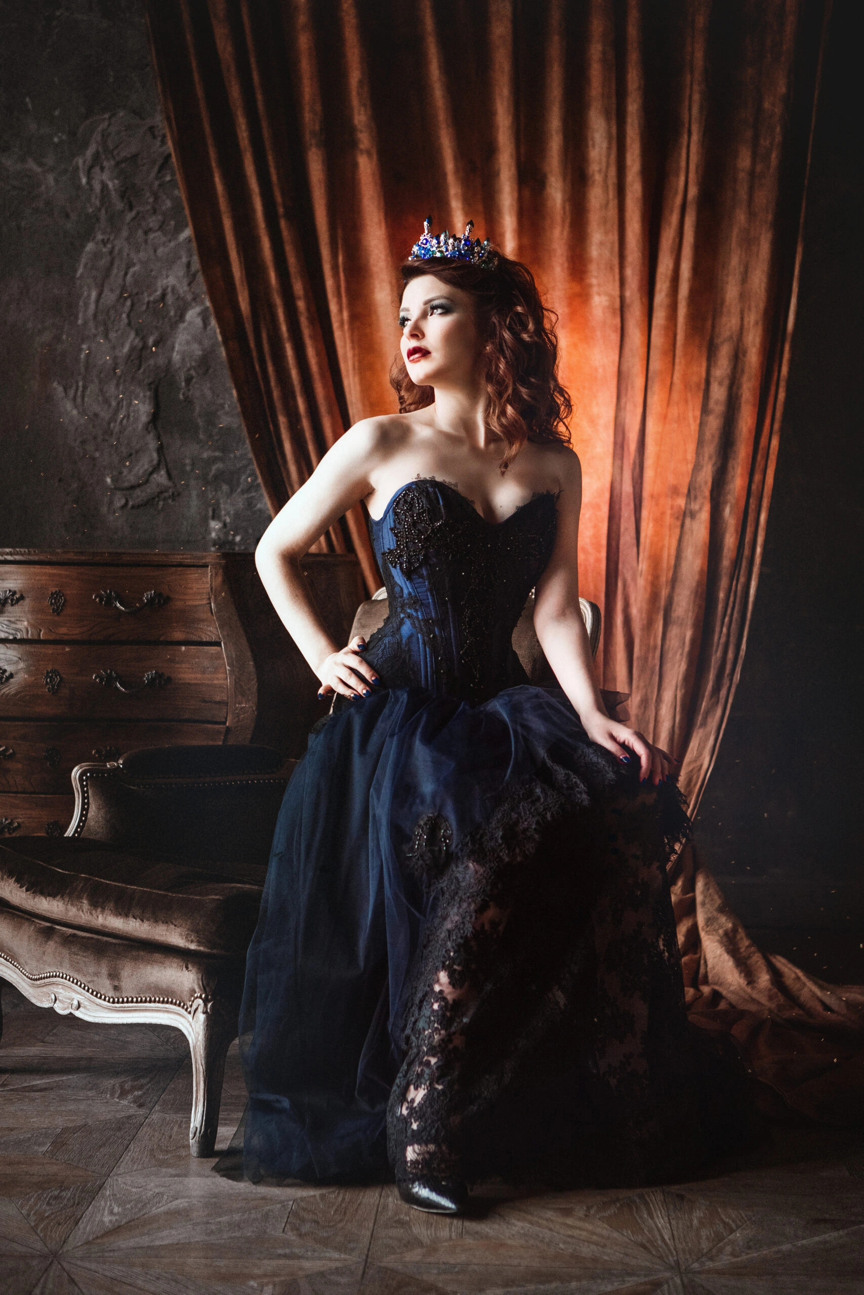 Woman in a blue over bust corset gown with a crown, sitting on a vintage chair with rich drapery in the background.
