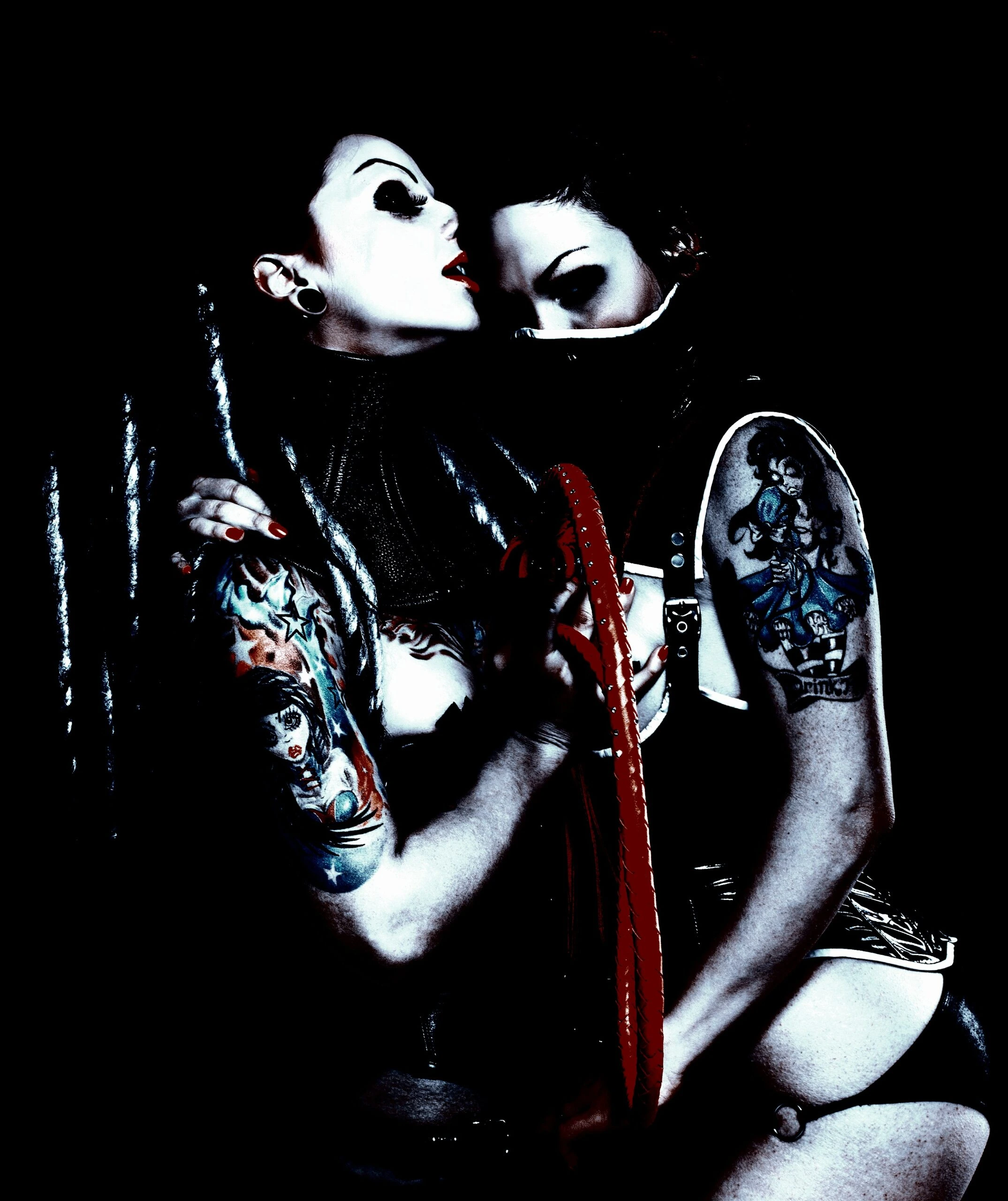 Two women with gothic tattoos, one wearing a mask and the other with bold makeup, in an intimate and darkly lit scene.