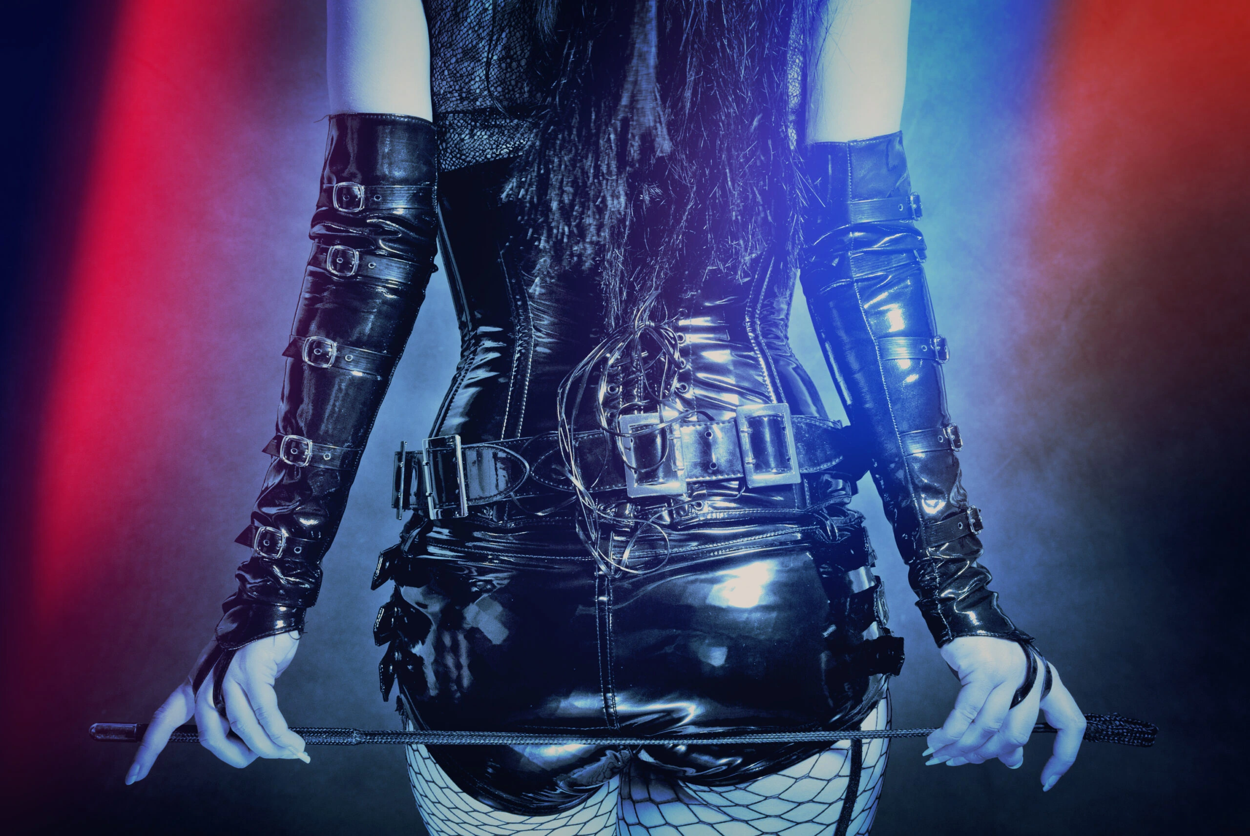 Back view of a woman in a black leather corset and gloves, holding a whip, highlighted with dramatic lighting.