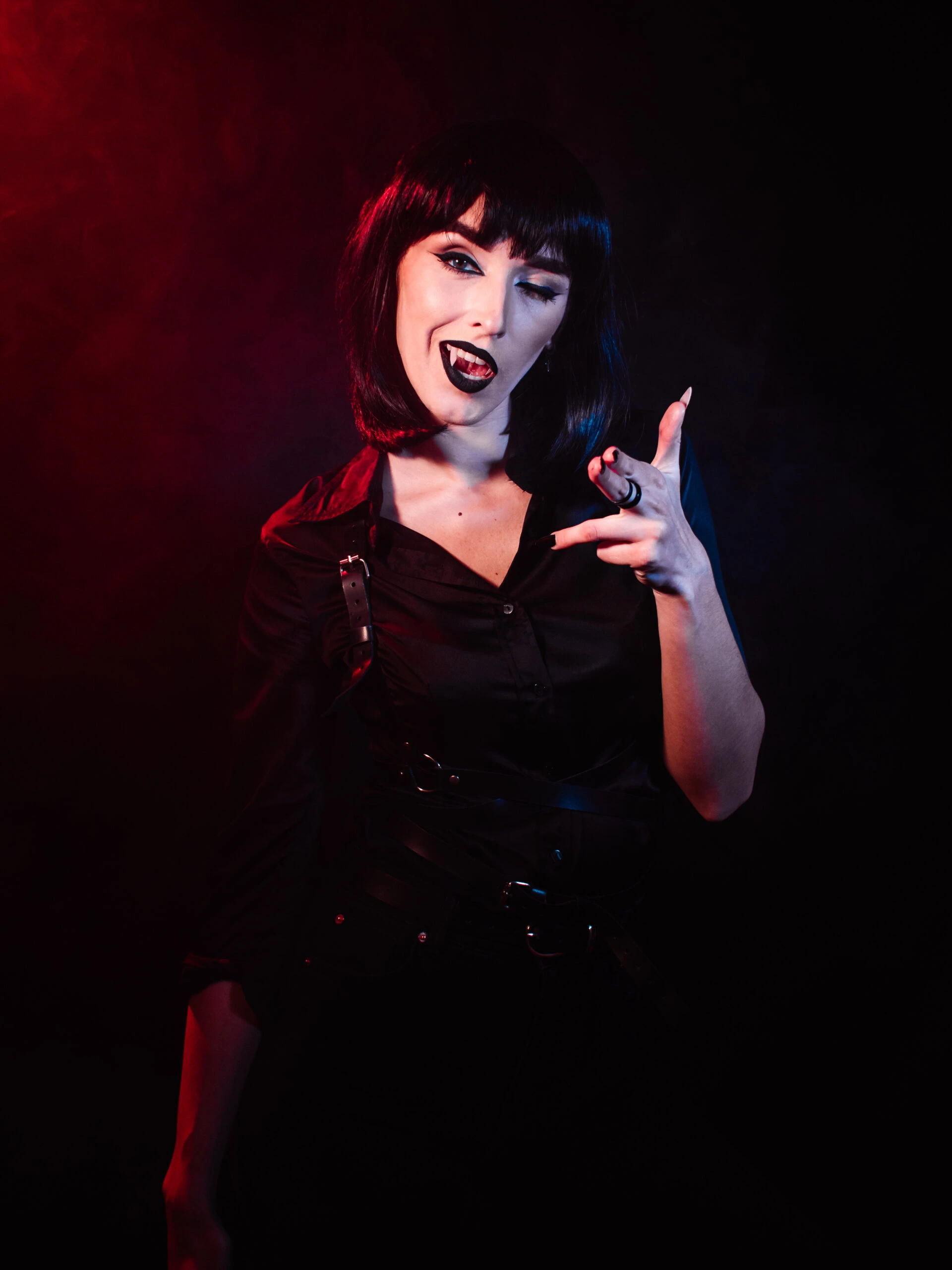 Woman with dark hair and Gothic makeup, wearing a black shirt, making a playful gesture in a dark setting.
