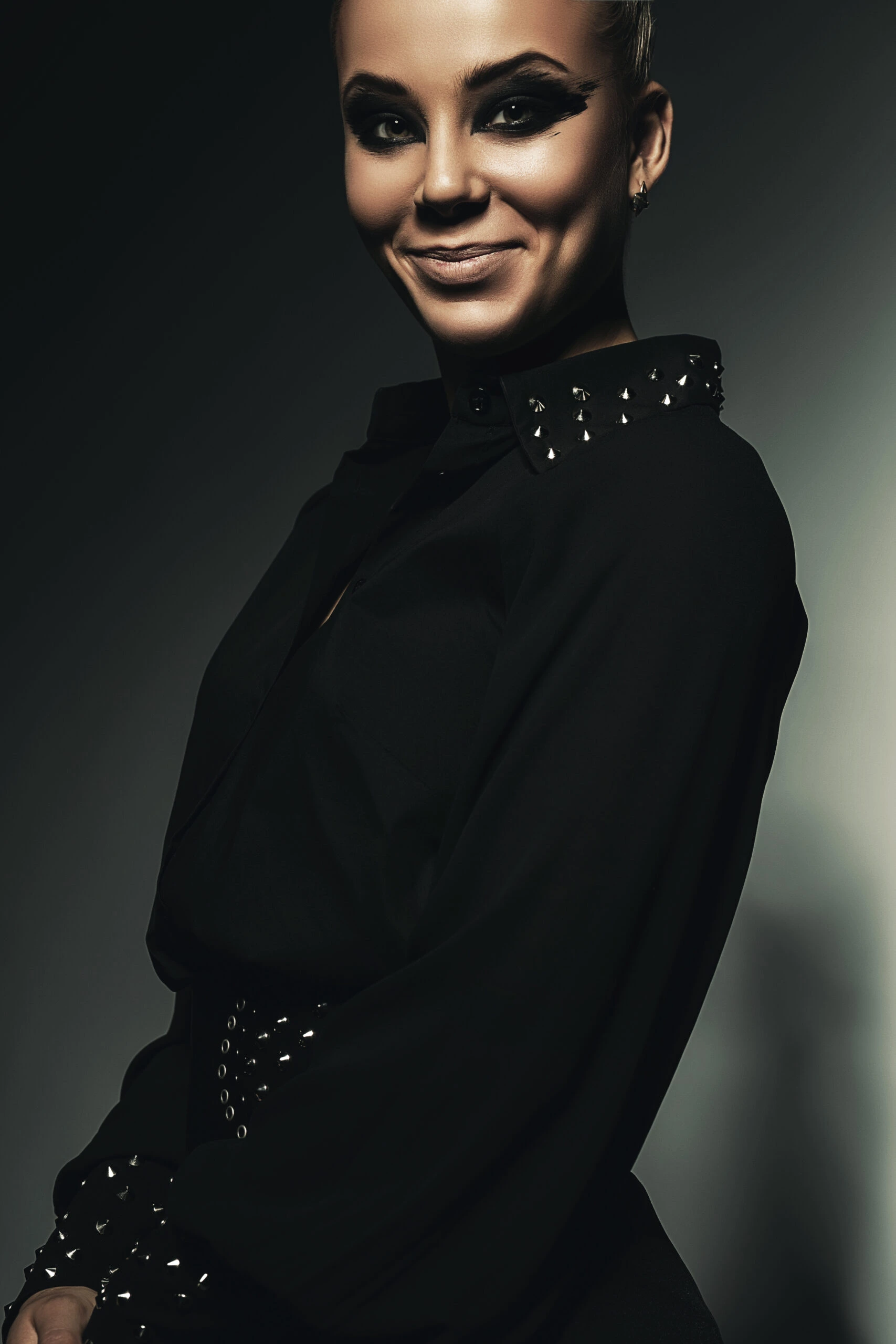 Woman in a dark Gothic shirt with studded collar, smiling with bold makeup.