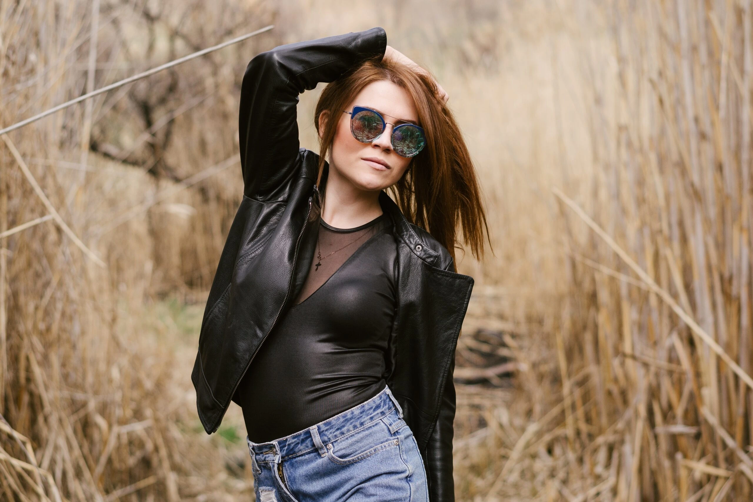 Woman in a leather jacket and sunglasses, posing confidently in a natural setting.