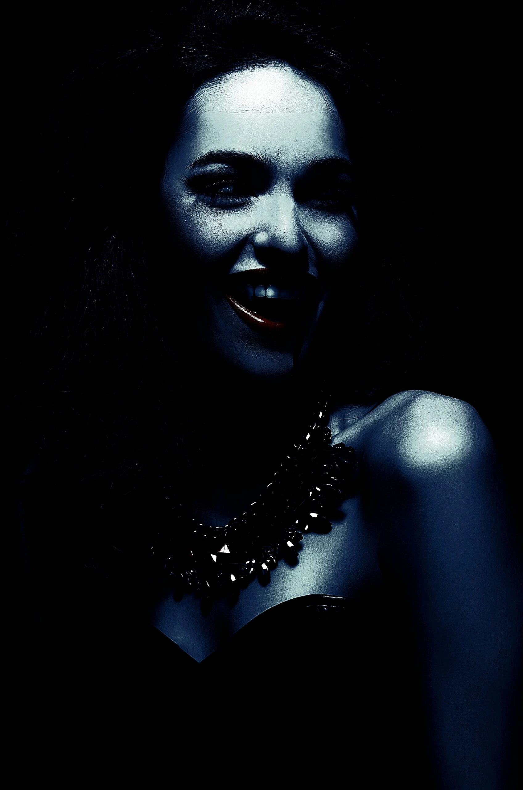 Woman with a dark, alluring smile, wearing a black strapless dress and statement necklace in vampire goth velvet attire.