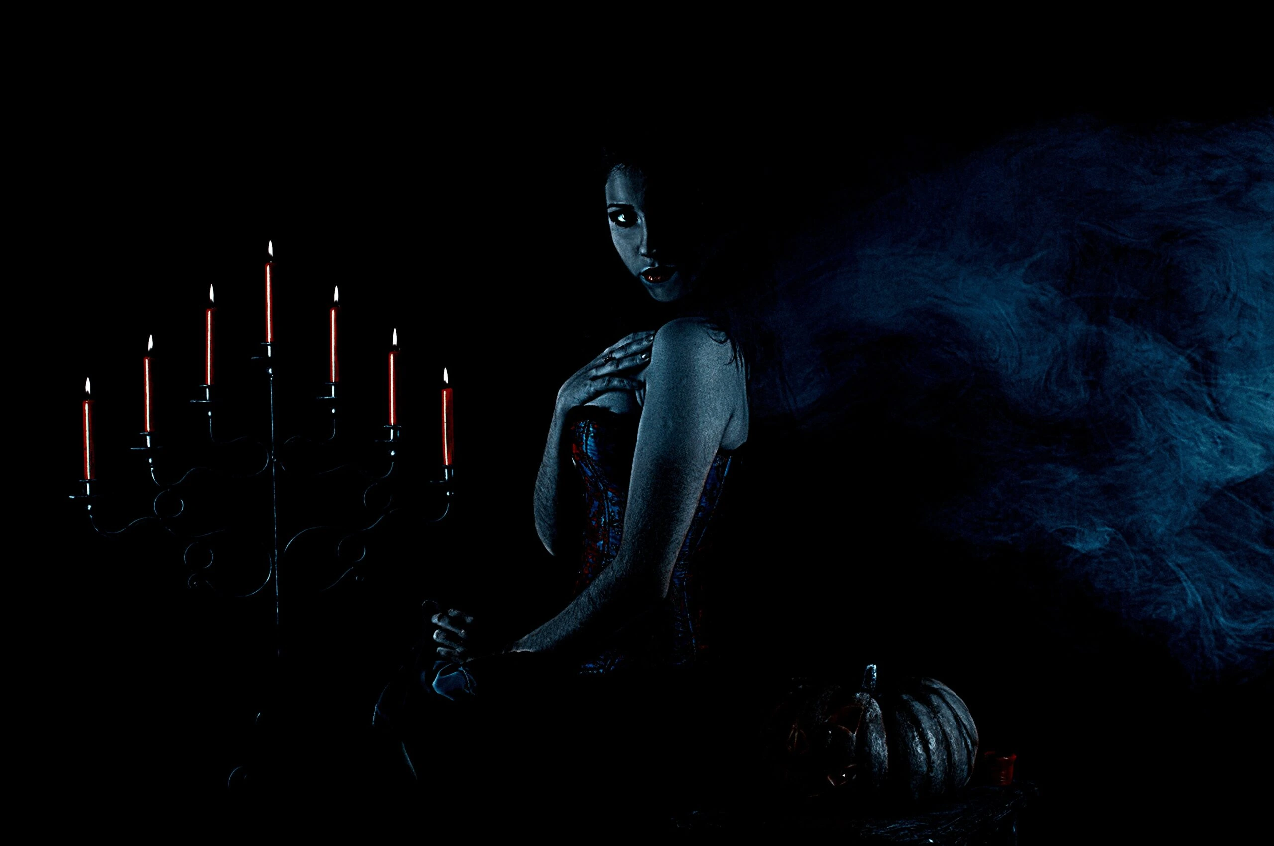 Gothic woman sitting next to a candelabra and pumpkin, wearing a corset and long skirt, in a dark, smoky setting.