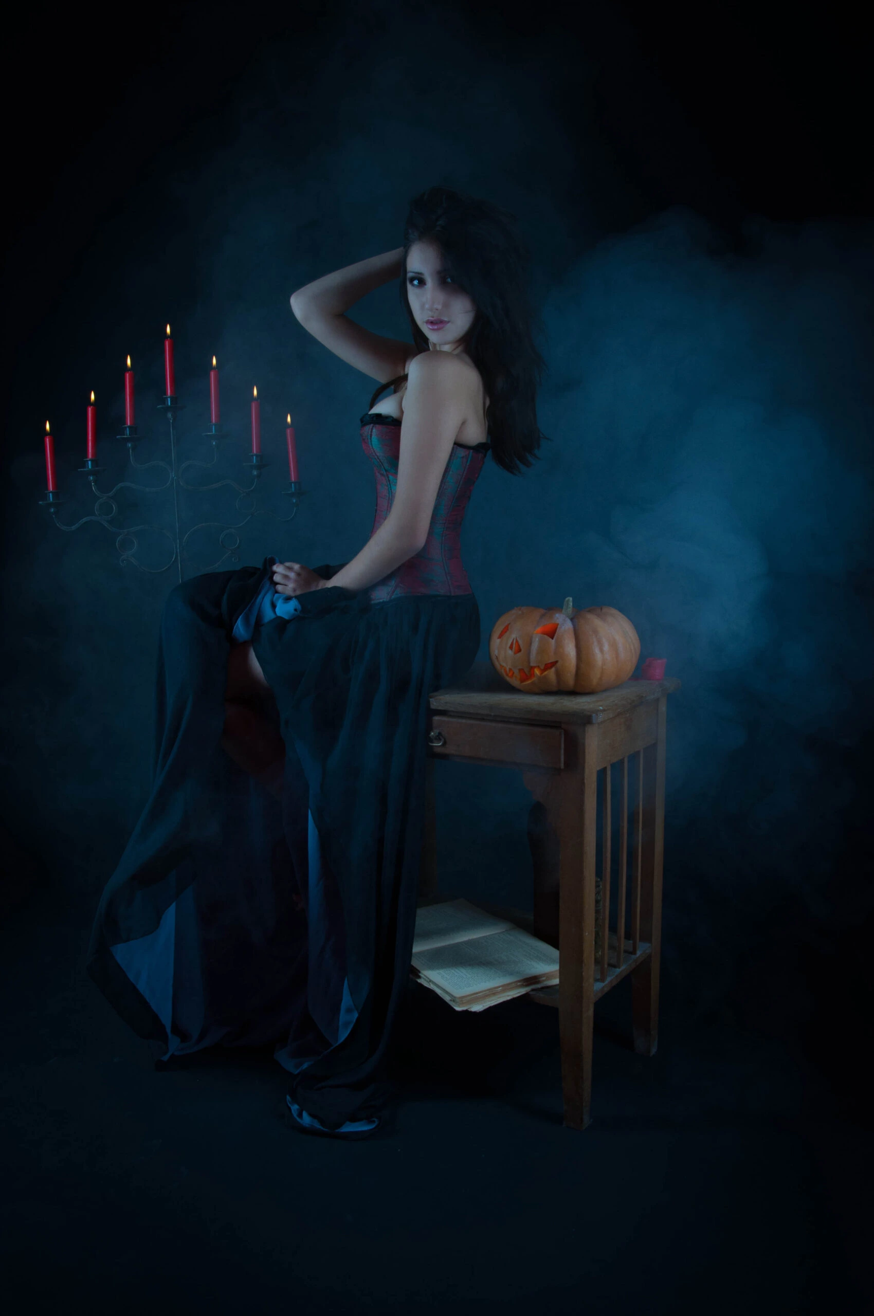 Gothic woman posing with a candelabra and pumpkin, wearing a corset and long skirt, surrounded by dramatic lighting and smoke.