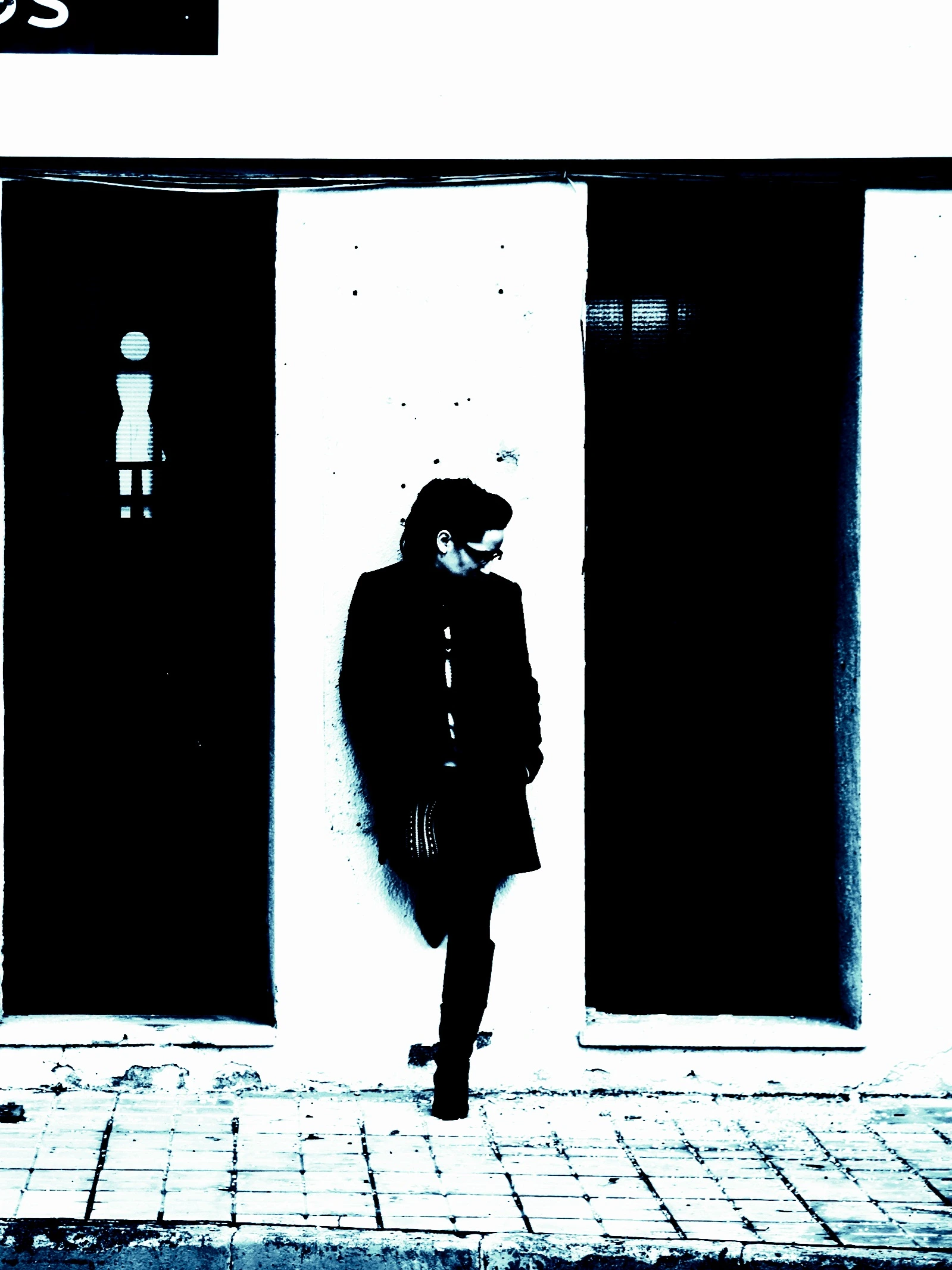 Person standing outside between restroom signs, wearing a long dark coat and looking away.