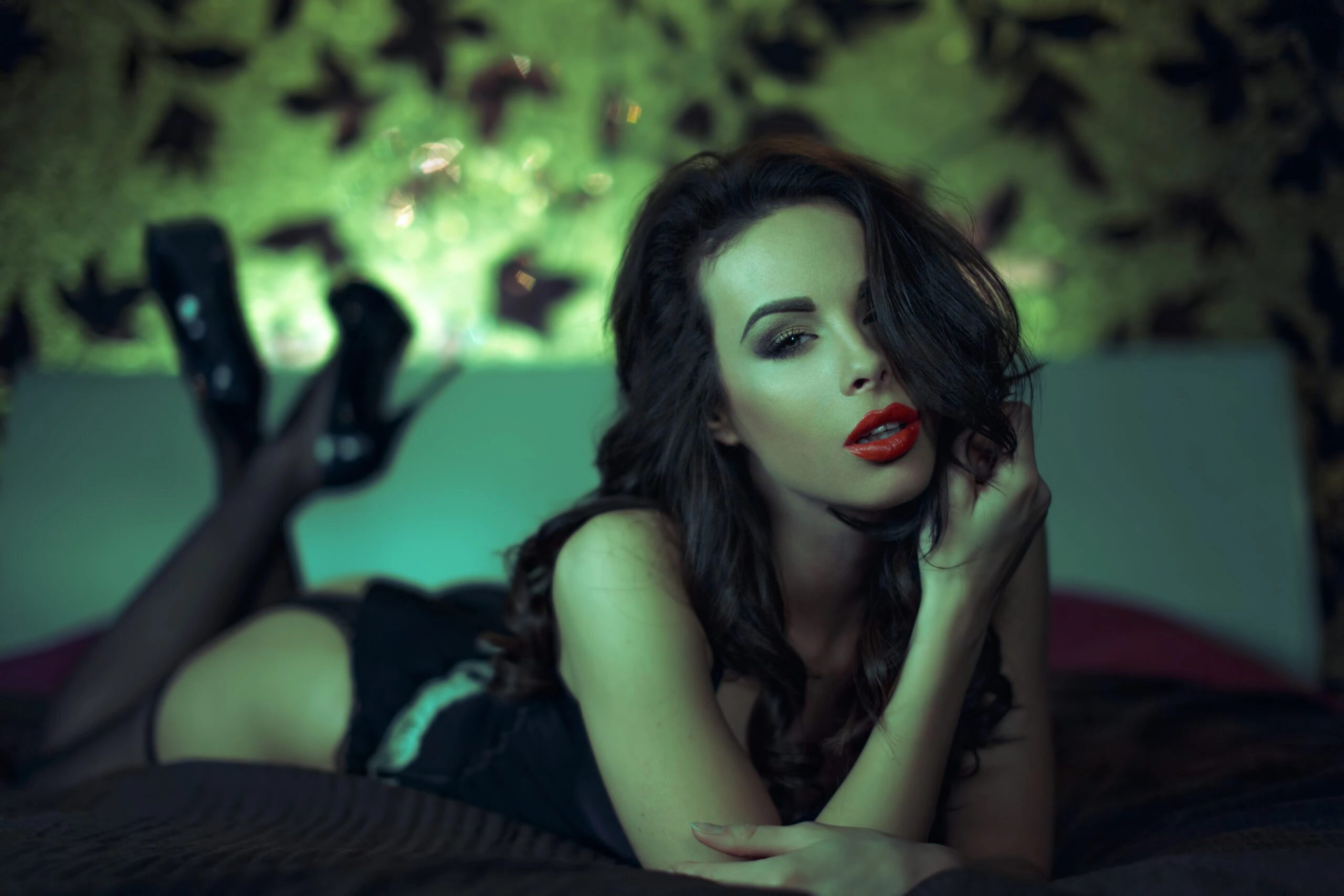 A woman in black lingerie and high heels lying seductively on a bed with a green and black patterned background, her red lips and tousled hair adding to the sultry mood.