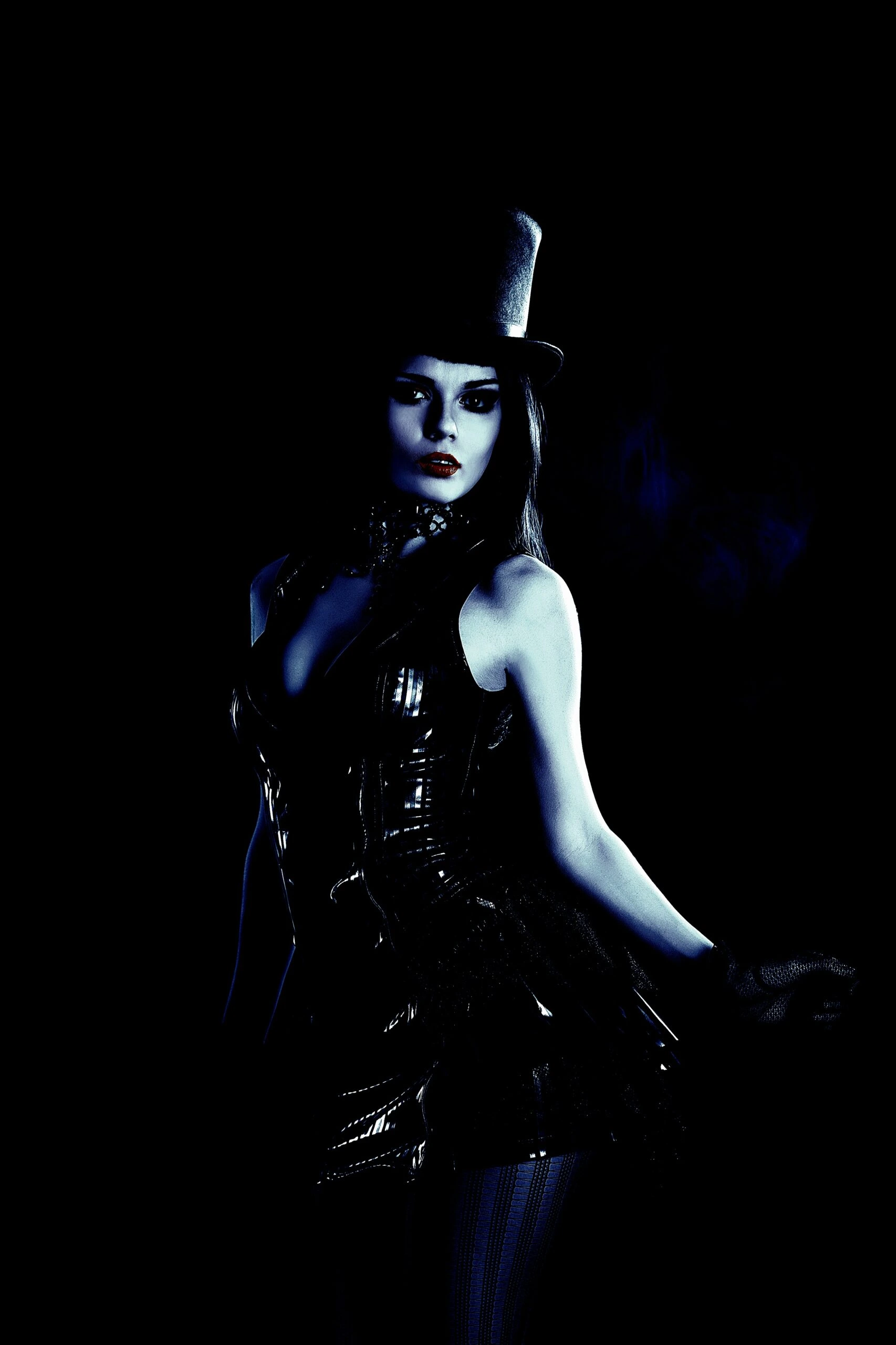 Gothic woman in a shiny black corset and top hat, with dramatic makeup and a dark background.
