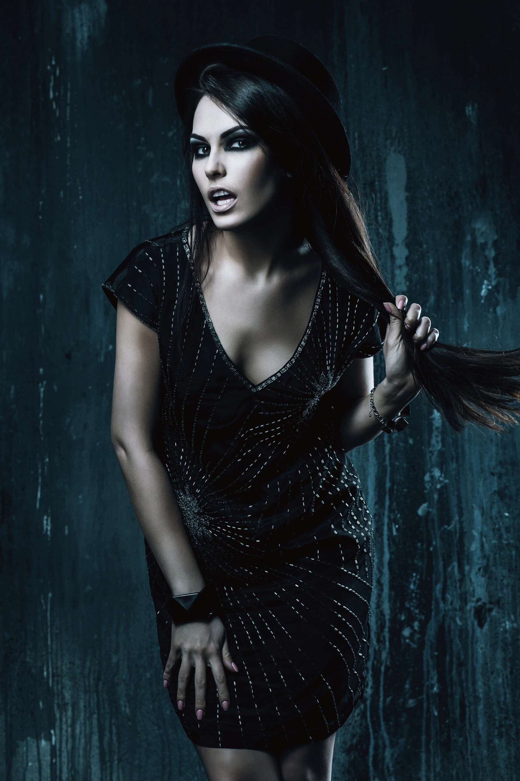Dark-haired woman in a short, sparkly gothic dress and hat, posing playfully with an intense gaze.