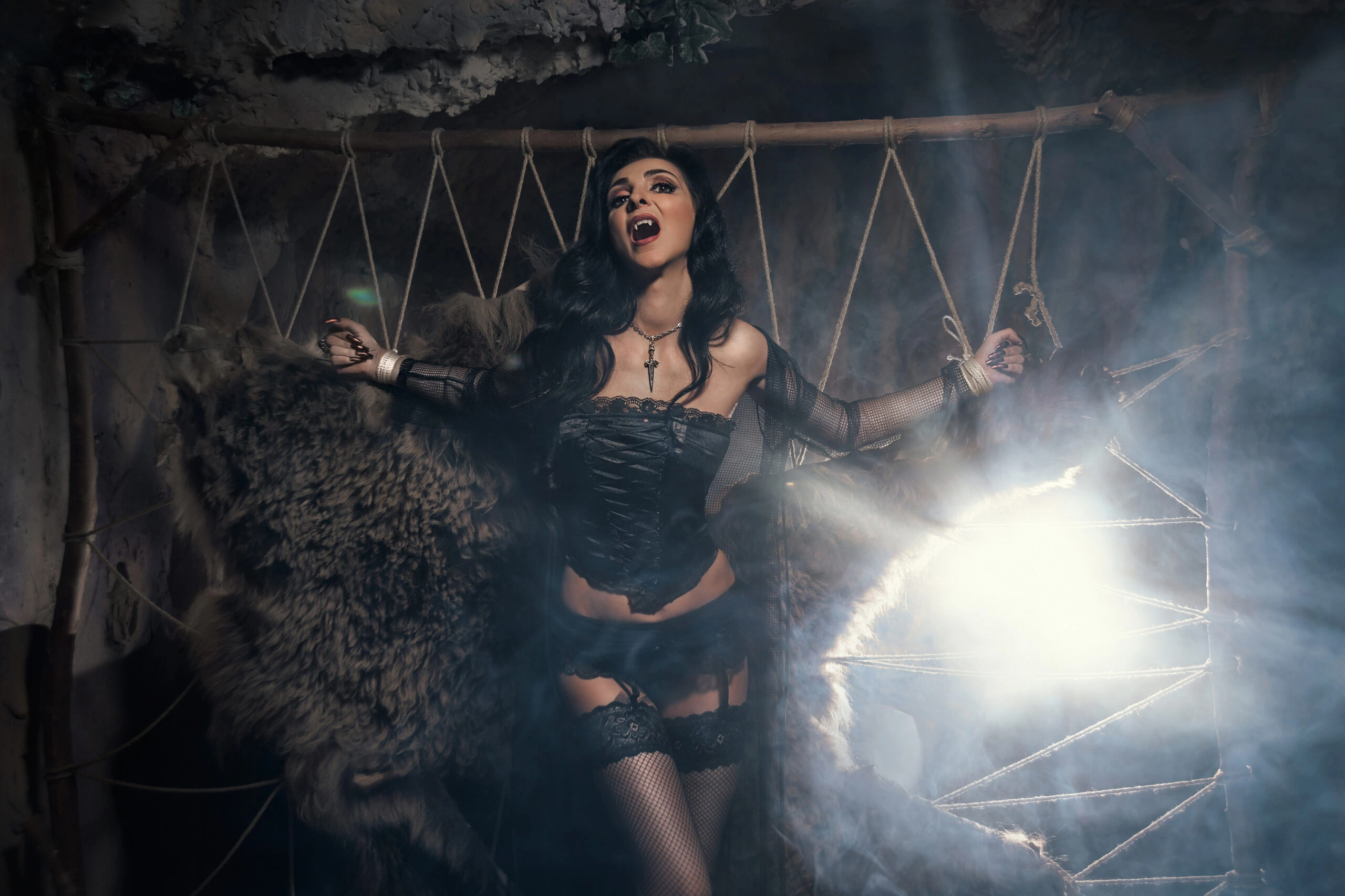 Gothic woman in a black corset and stockings, posed dramatically in a cave setting with fur and mist.