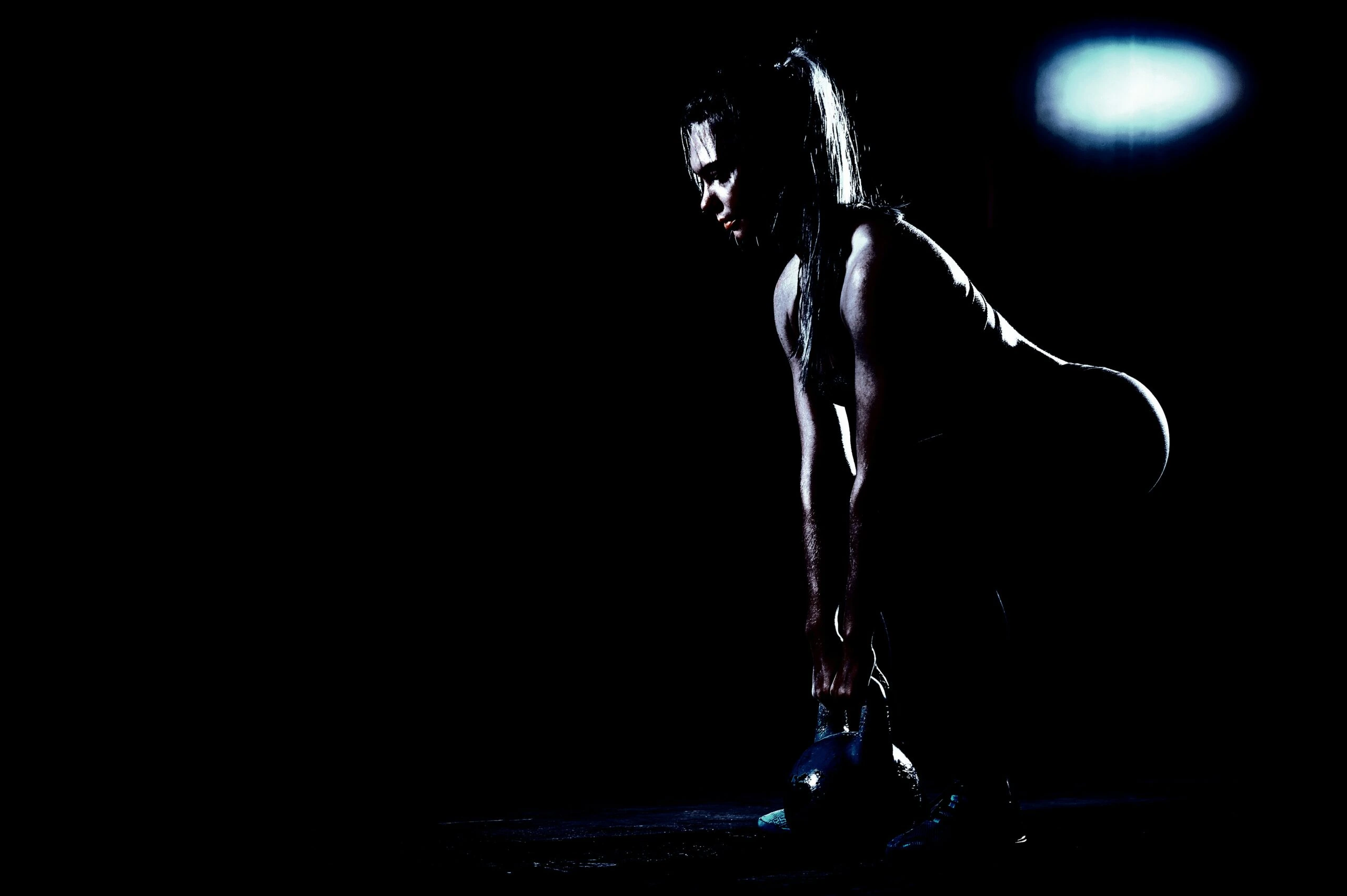 Woman performing a kettlebell deadlift in a dark gym, her muscles highlighted by dramatic lighting.