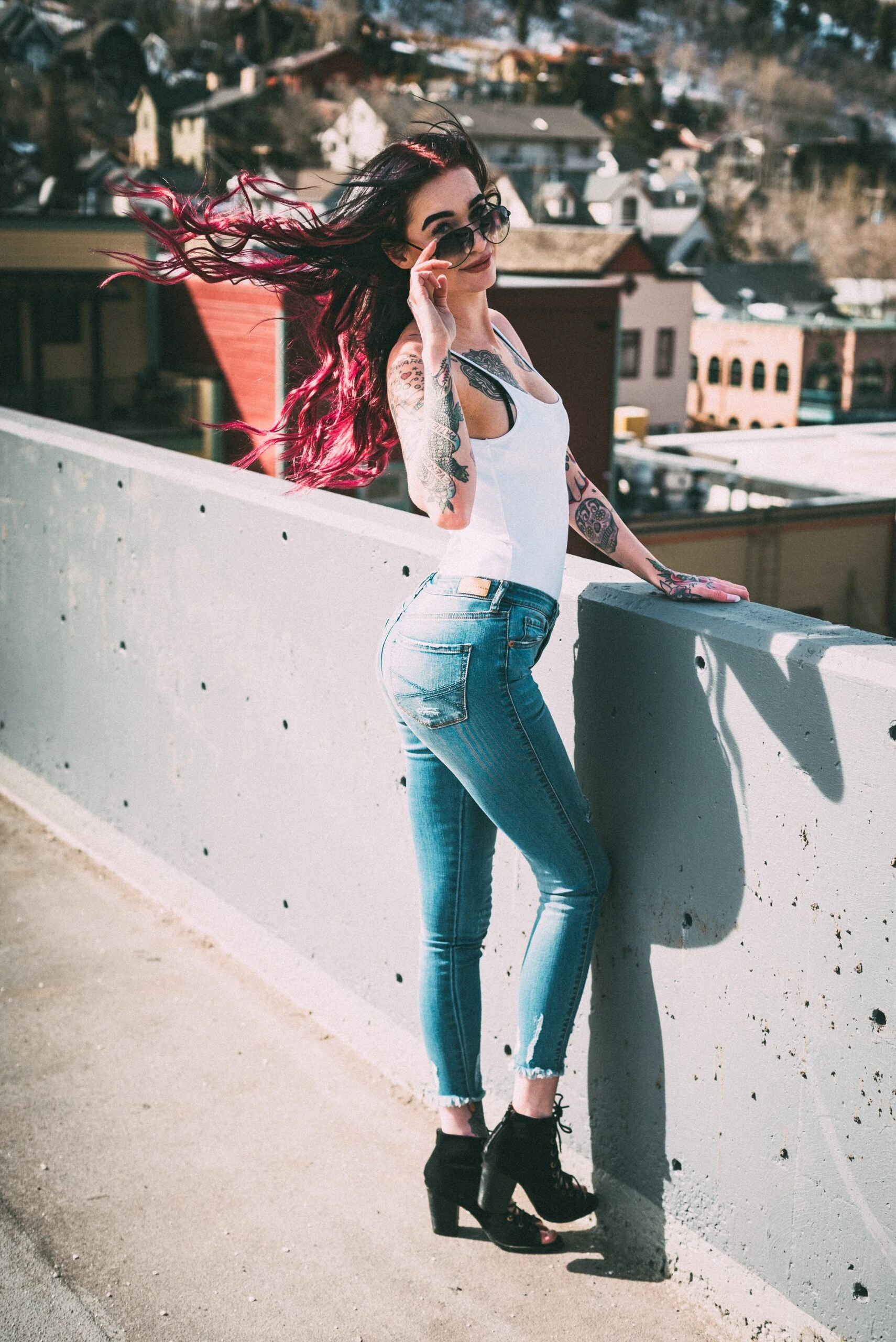 High-contrast image of a woman with long dark hair and red highlights, wearing a white tank top and jeans, posing with aviator sunglasses on a rooftop.