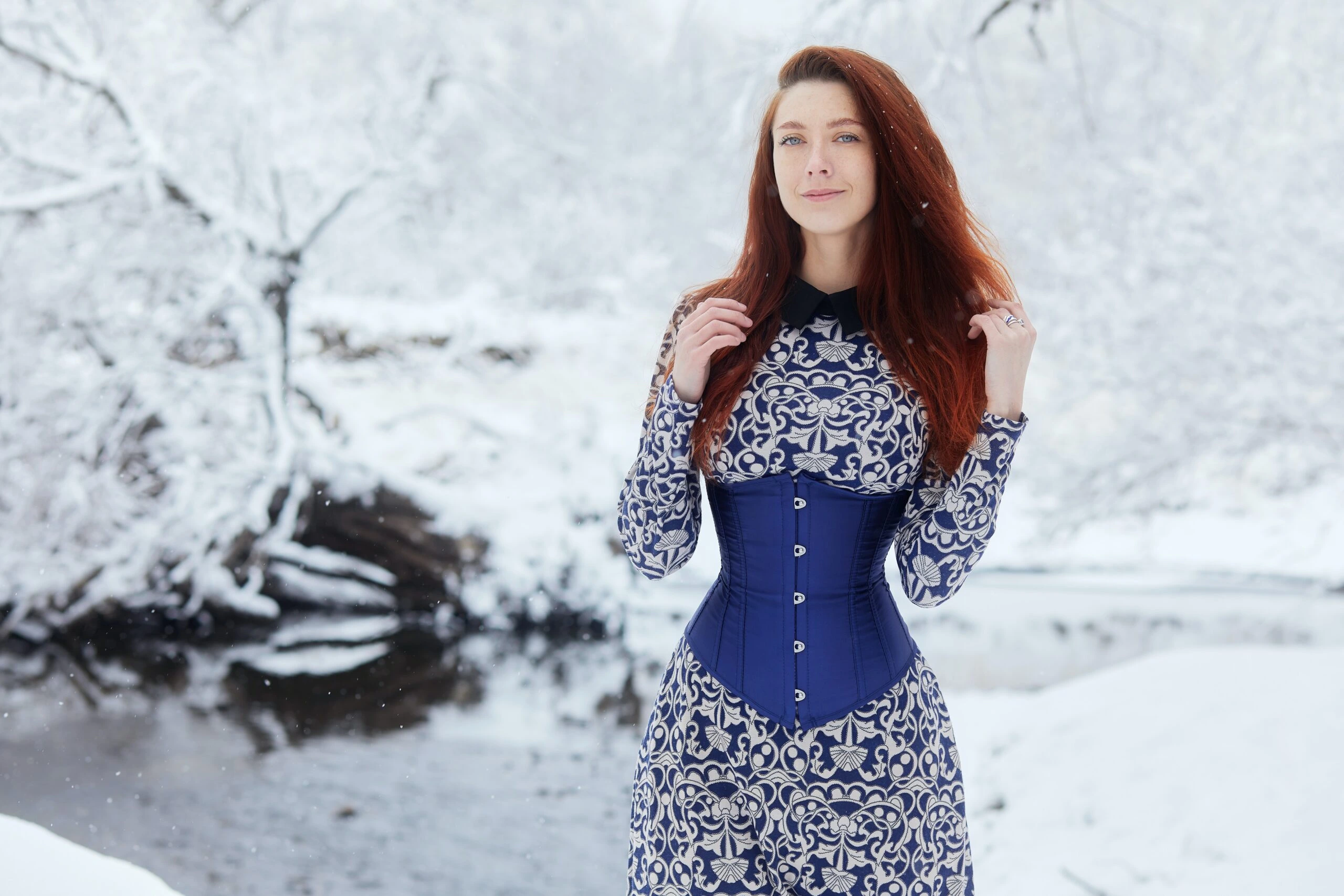 Woman in a blue corset and patterned dress standing in a snowy landscape, showcasing winter corset fashion.