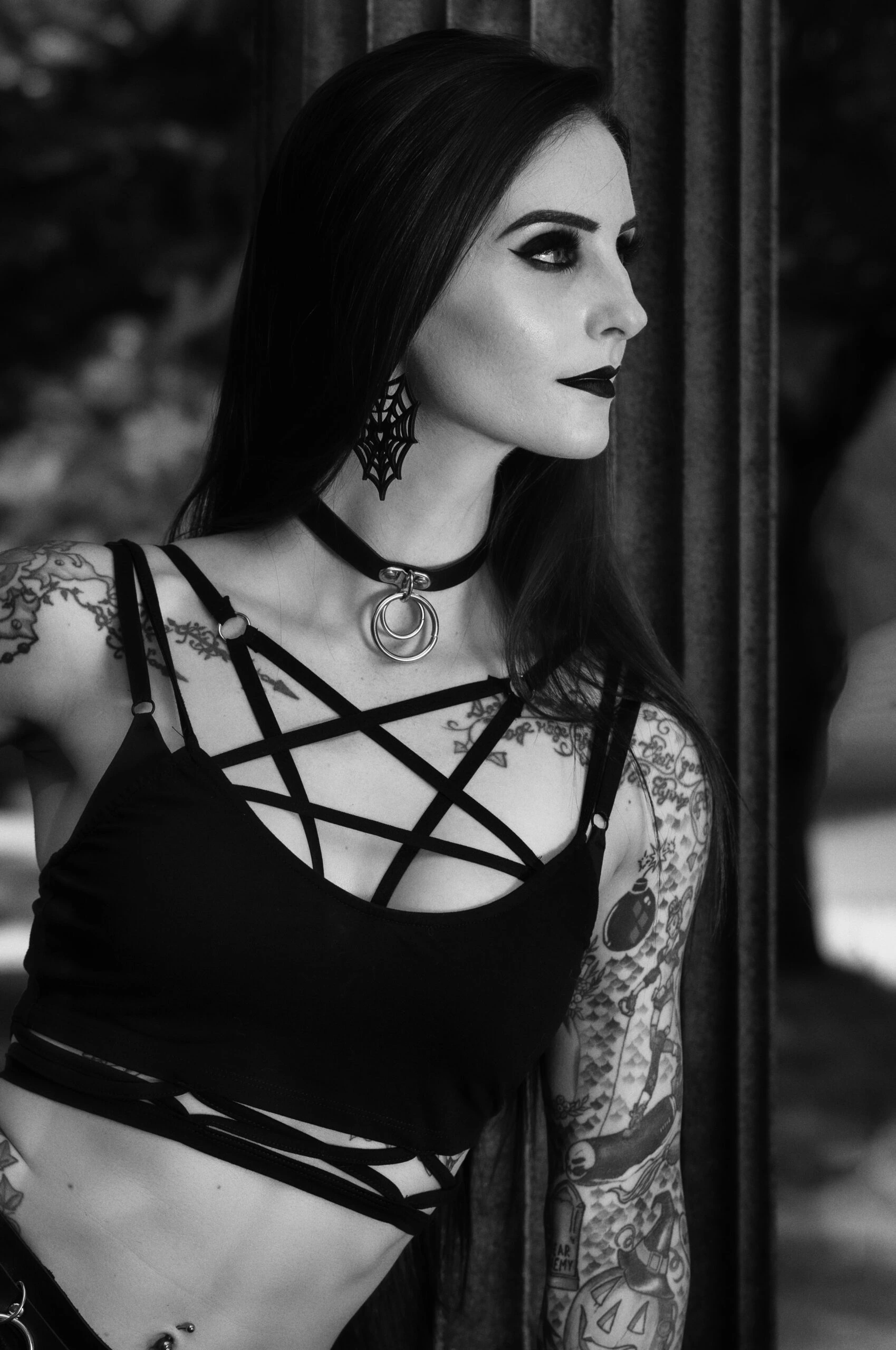 Gothic woman with black hair and dark makeup, wearing a dramatic choker necklace with red gems, standing against a backdrop of lit candles.