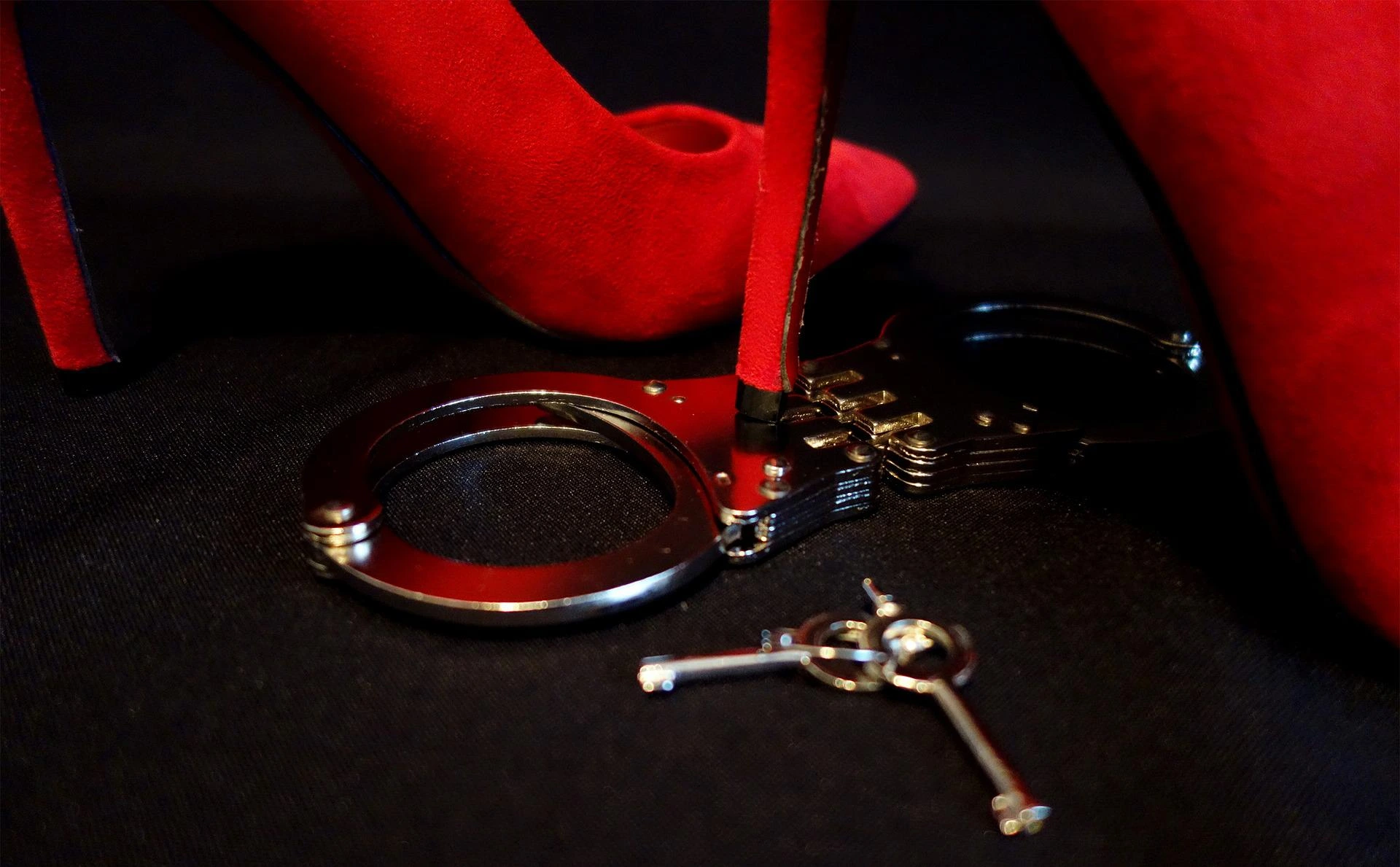 Red high heel resting next to a pair of handcuffs and keys on a black surface.