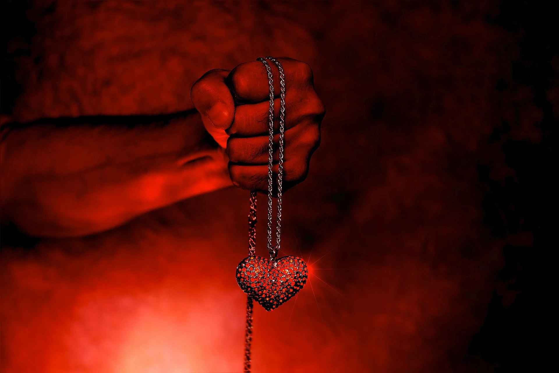 A hand clenched around a chain holding a red heart-shaped pendant against a dark, red-lit background.