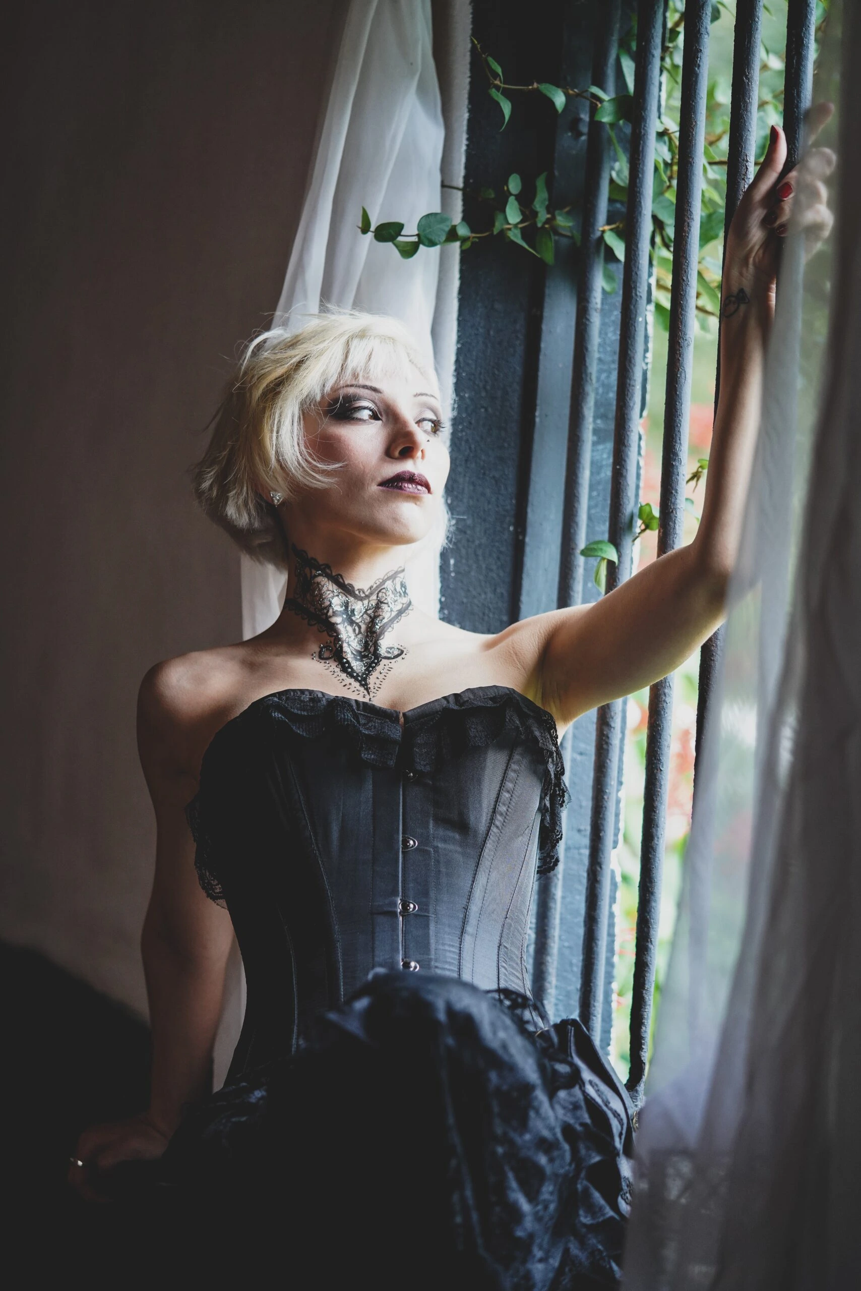 A gothic woman in a black overbust corset with short platinum hair, gazing out a window with an ethereal expression.