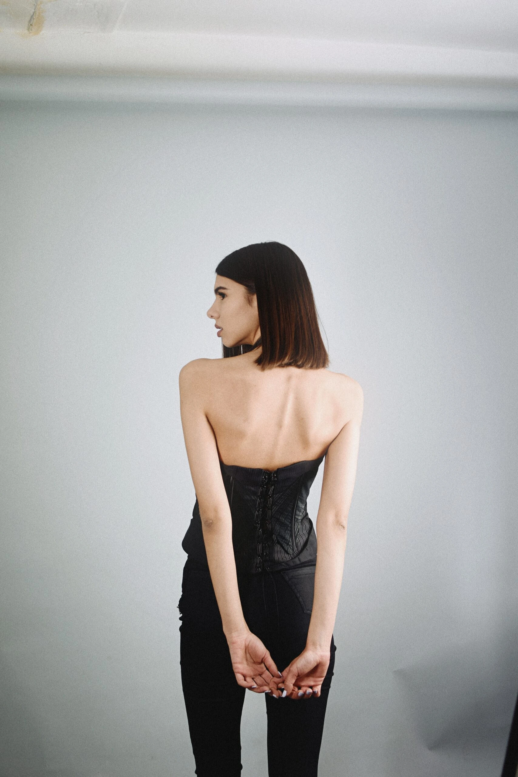 A model in a black corset with a minimalist backdrop, standing with her back turned and arms clasped, highlighting the corset’s design and fit.