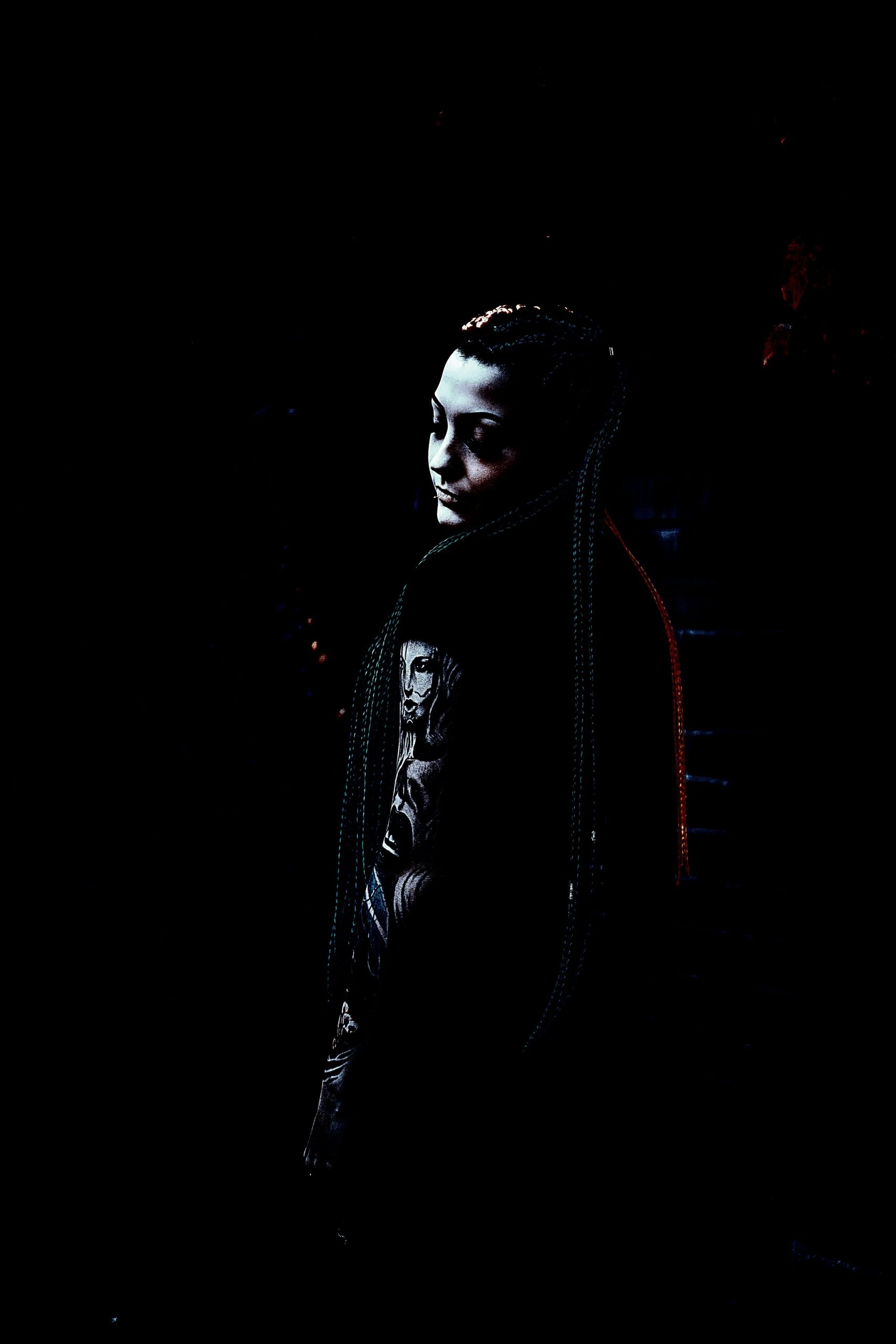 Gothic woman with long braids and intricate tattoos standing in a shadowy environment. 