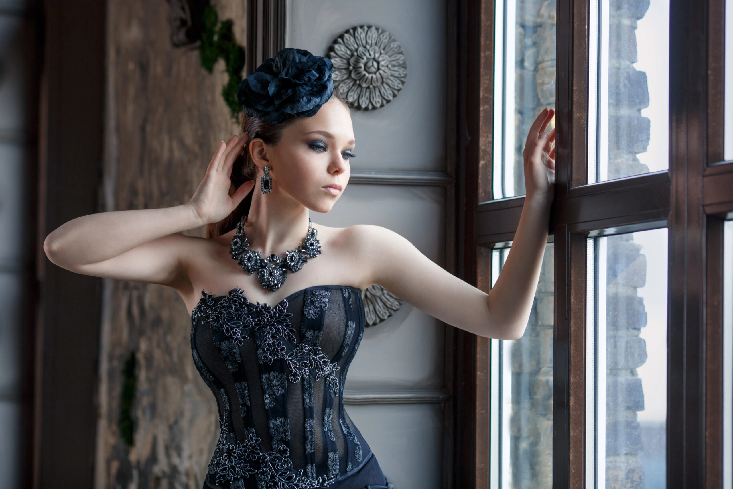Elegant woman in a Victorian-inspired gothic outfit, featuring a black corset and intricate jewelry, gazing out of a window in a historic setting.