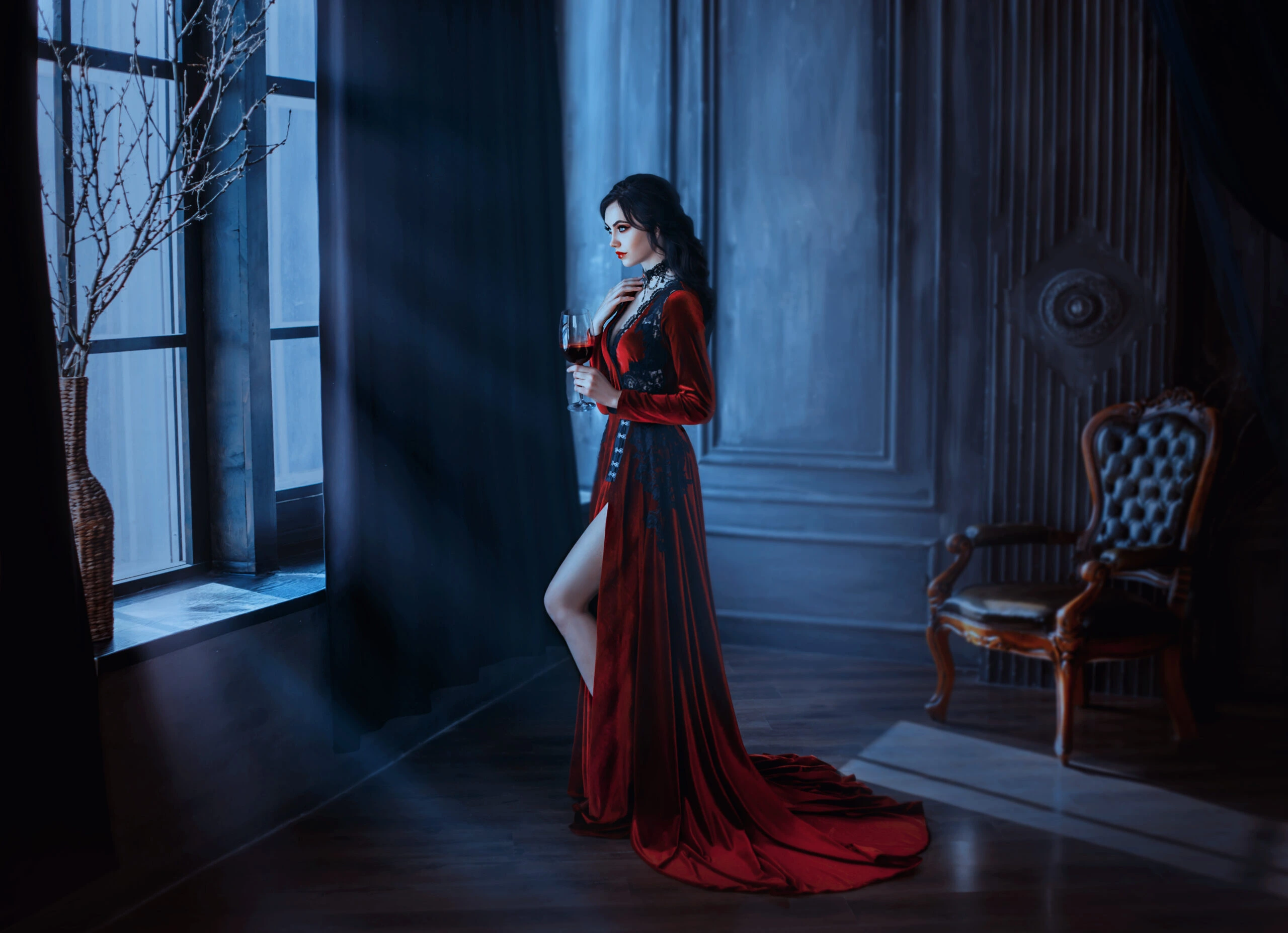 A woman dressed in a long, elegant red velvet gown with lace detailing, standing near a window in a dark gothic room, holding a glass of red wine.