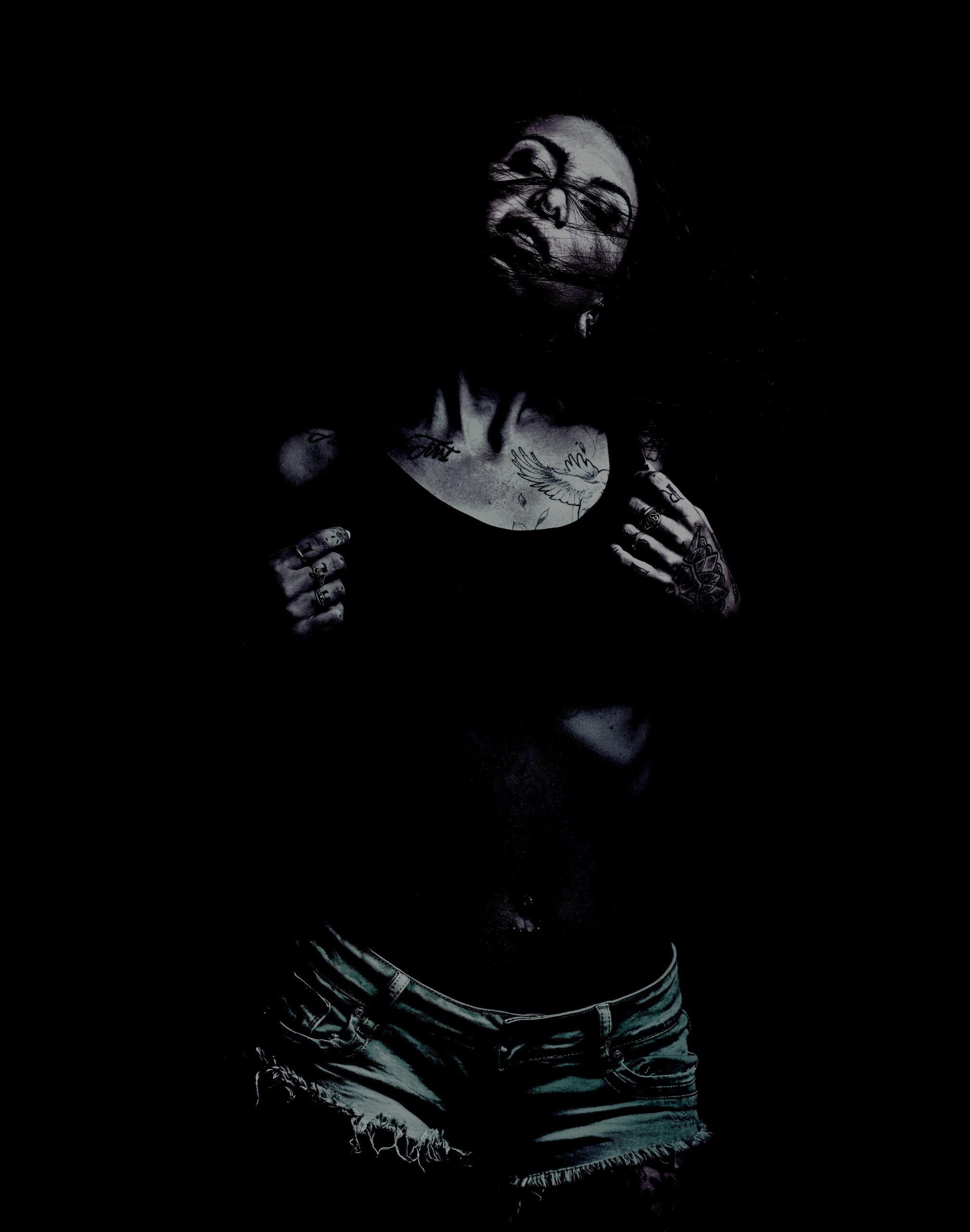 Woman with tattoos and piercings posing in a dark, moody setting, wearing denim shorts.