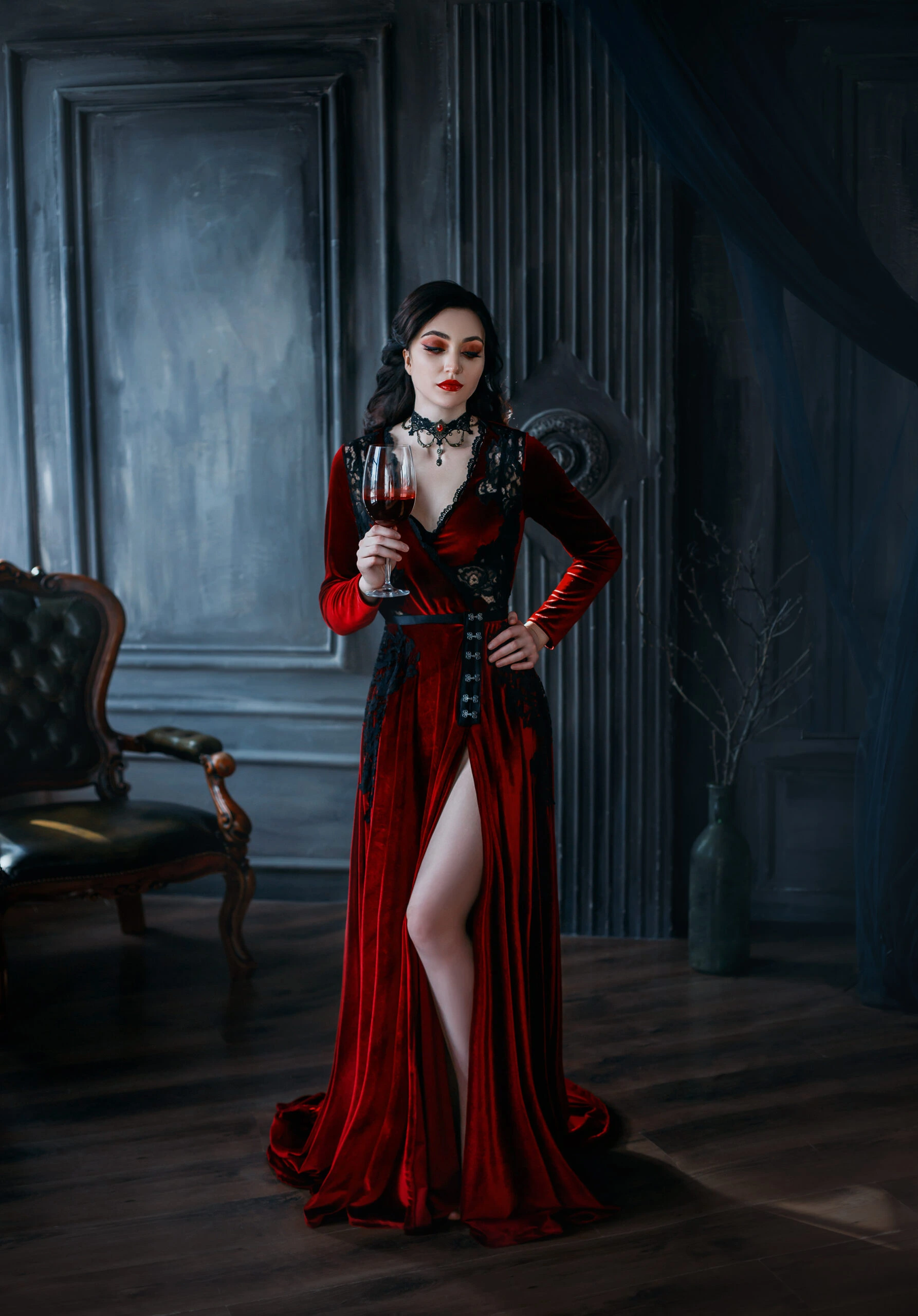A woman in a full-length red velvet gothic gown with a thigh-high slit, standing in a luxurious dark room, holding a glass of red wine.