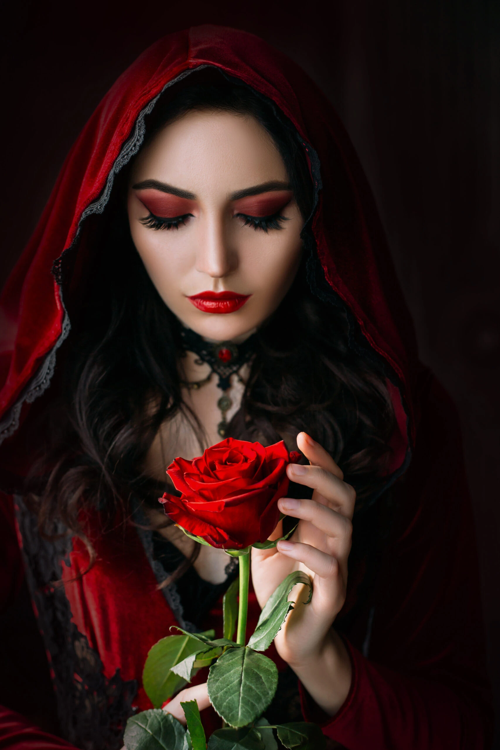 A woman wearing a red hooded cloak, holding a vibrant red rose, her eyes closed and makeup emphasizing her pale skin and red lips, evoking a gothic fairy tale vibe.