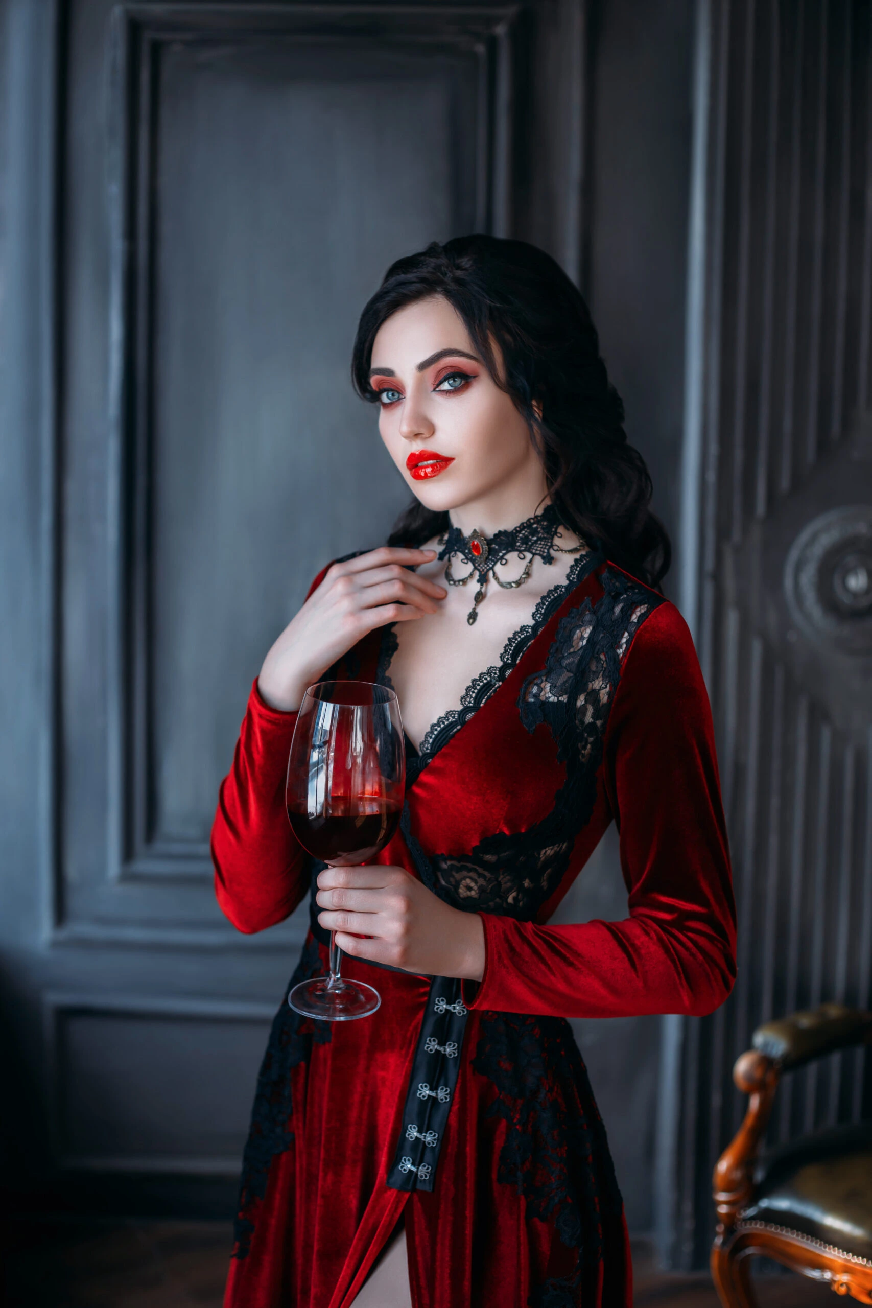 A woman dressed in a seductive red velvet dress with black lace details, holding a glass of red wine, standing confidently in a gothic-themed room.