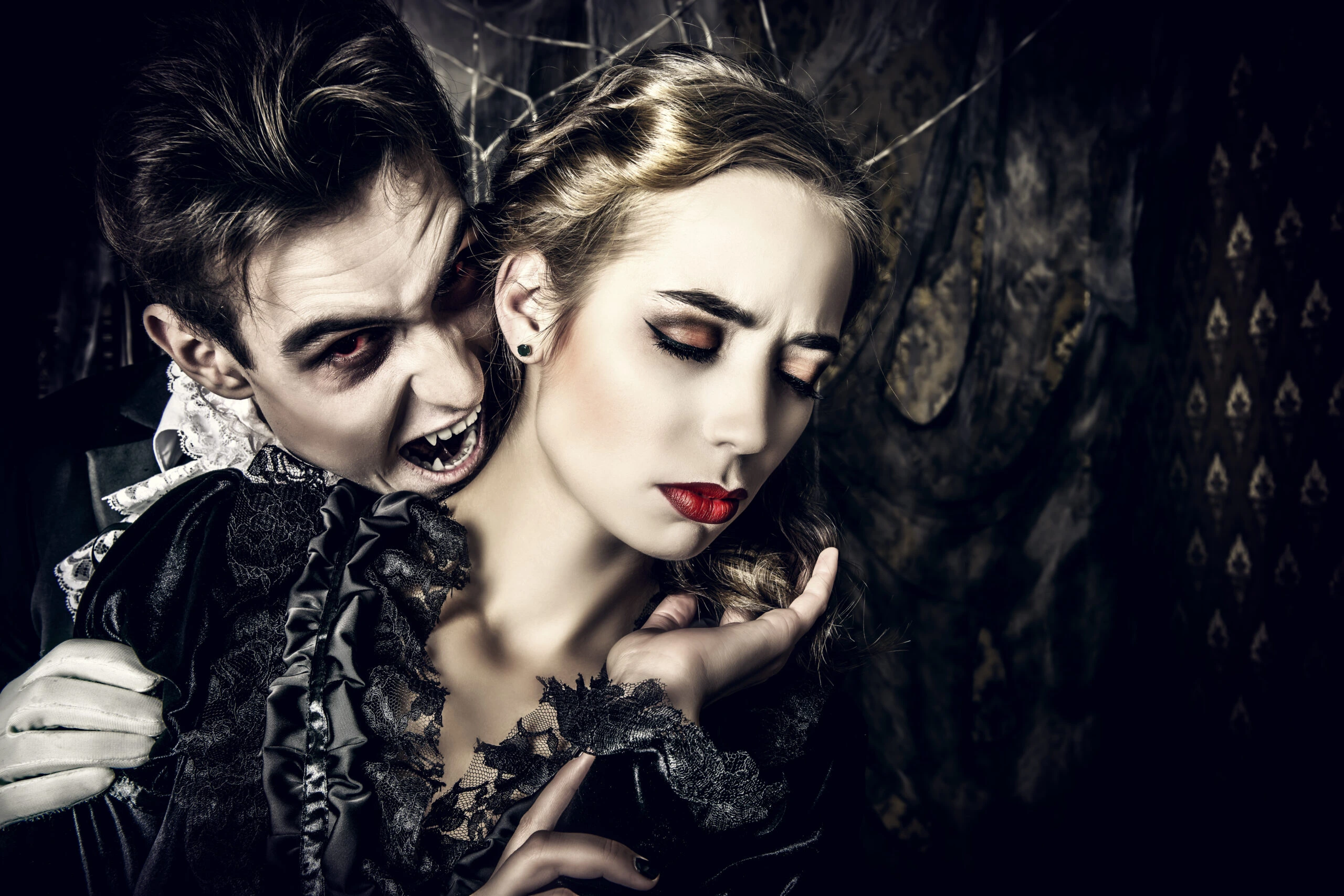 A male vampire with fangs biting a woman's neck, both dressed in gothic attire with dark, dramatic makeup in a haunting atmosphere.