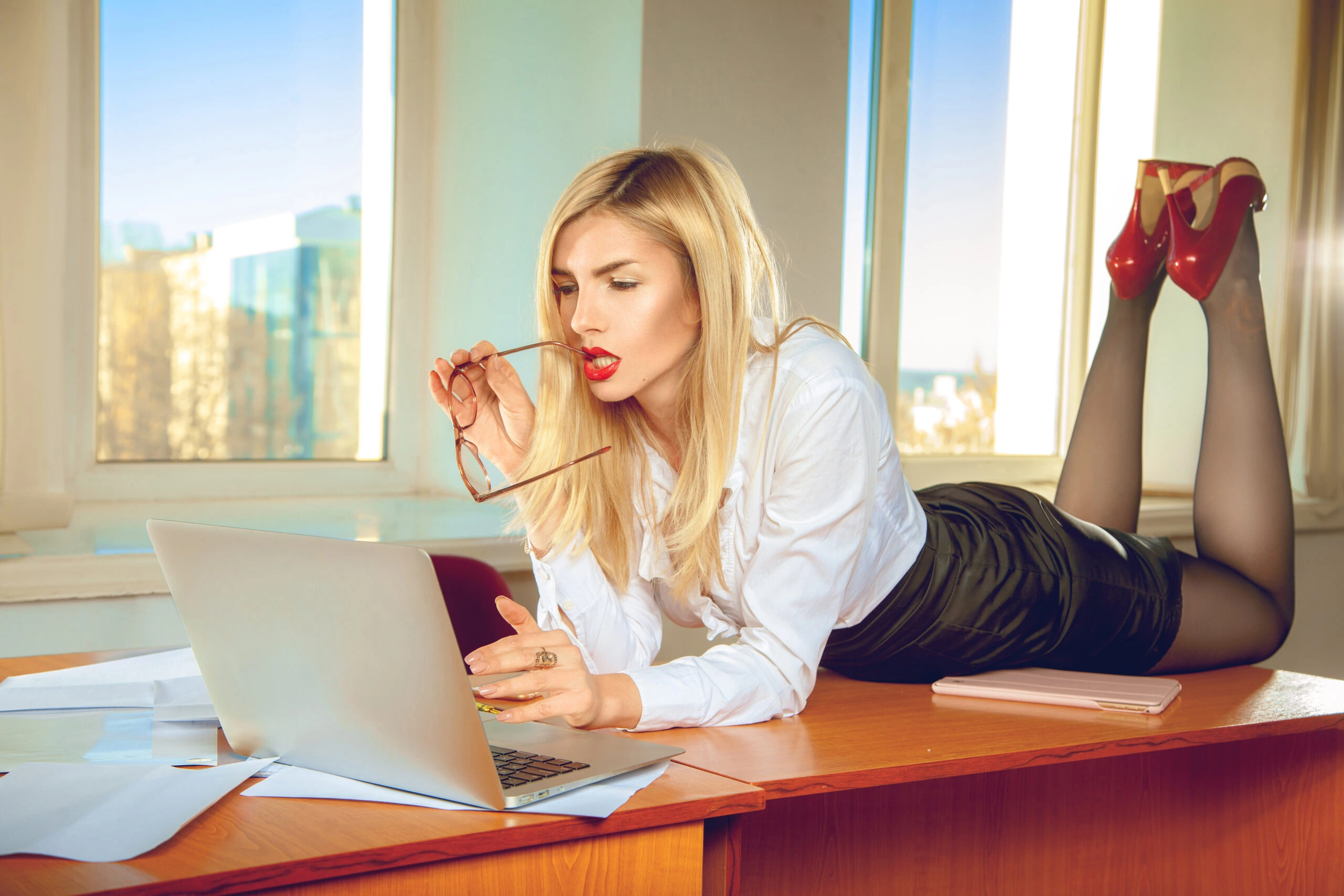 Stylish businesswoman in red heels and stockings, lying on a desk and working on a laptop.