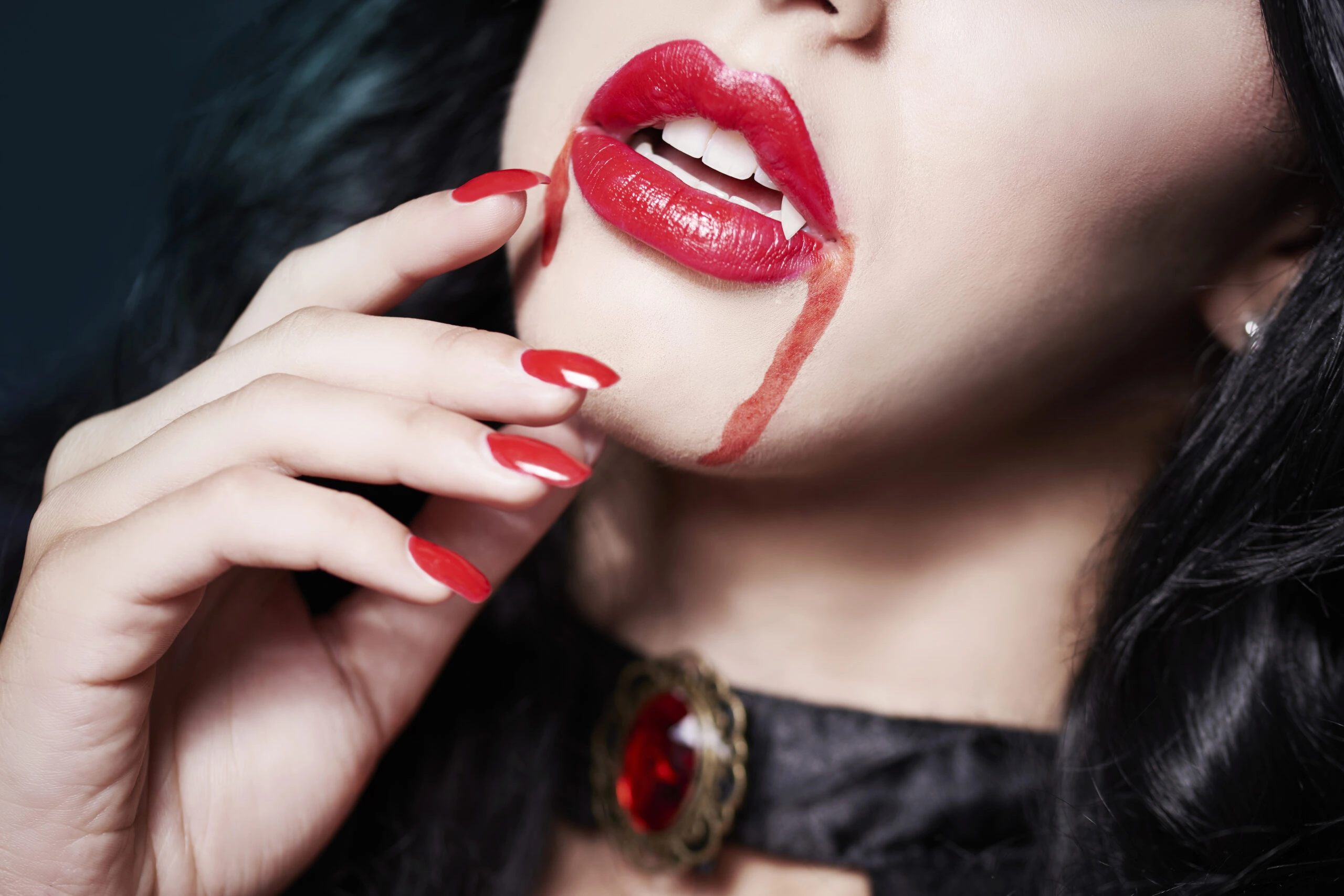 Close-up of a woman’s lips and red nails, with vampire fangs visible and blood dripping from her mouth, evoking a seductive vampire aesthetic.