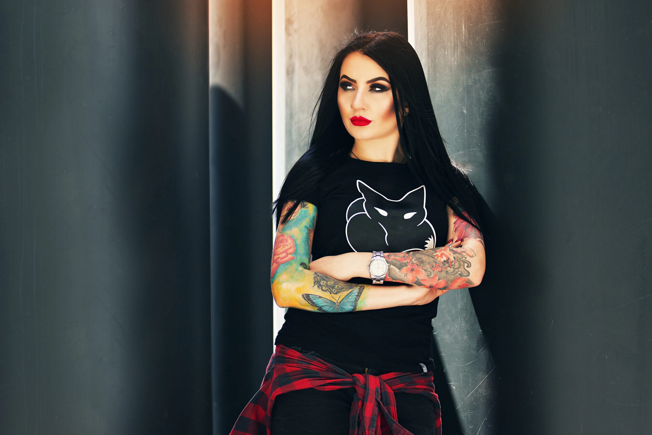 Woman with long black hair, wearing a black t-shirt with a fox graphic, colorful tattoos on both arms, standing with arms crossed and leaning against a dark pillar, exuding a bold, goth-inspired look.