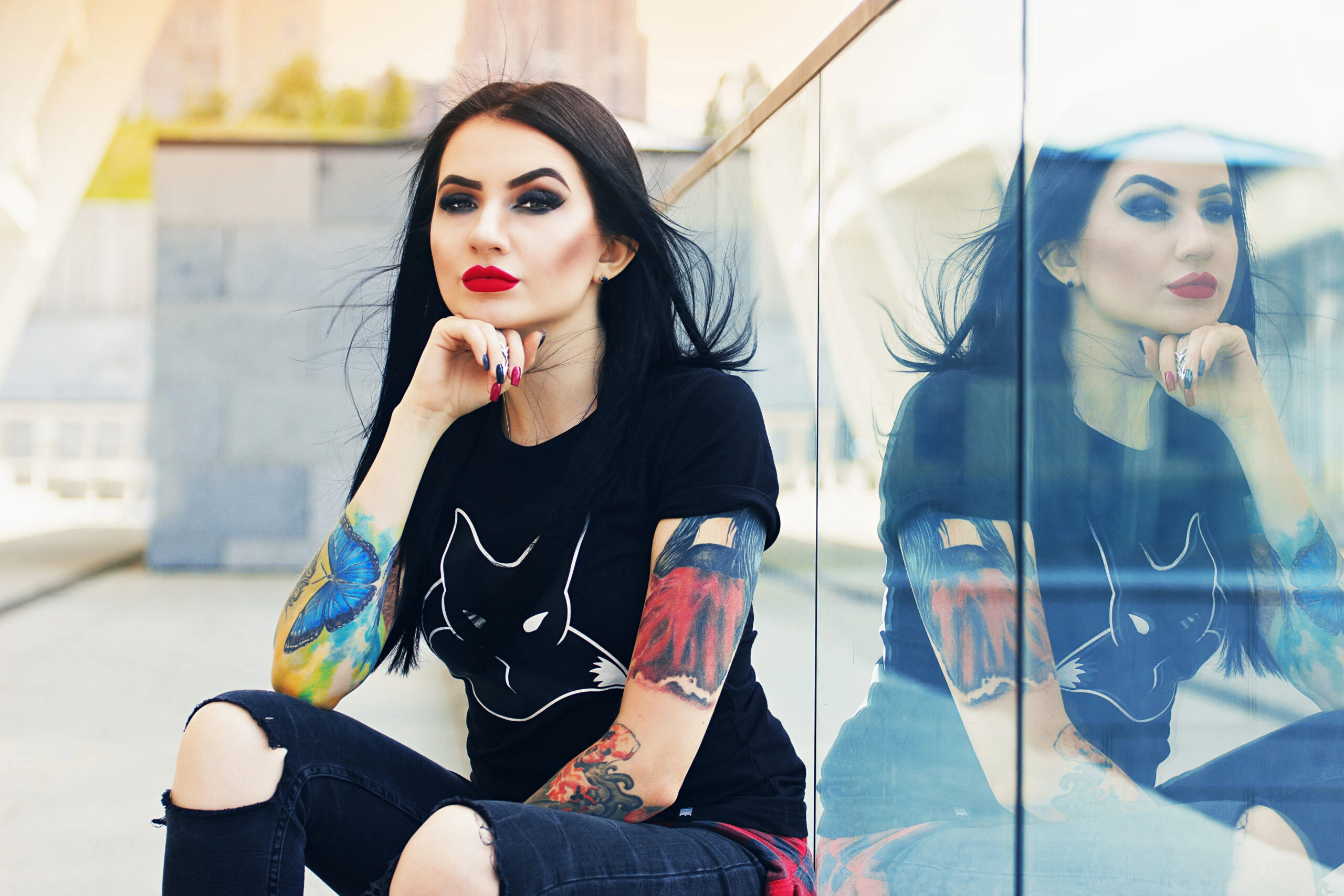 Woman with black hair, bold red lipstick, wearing a black t-shirt with a fox graphic, seated against a reflective glass wall, showcasing her tattoos and goth-inspired fashion.