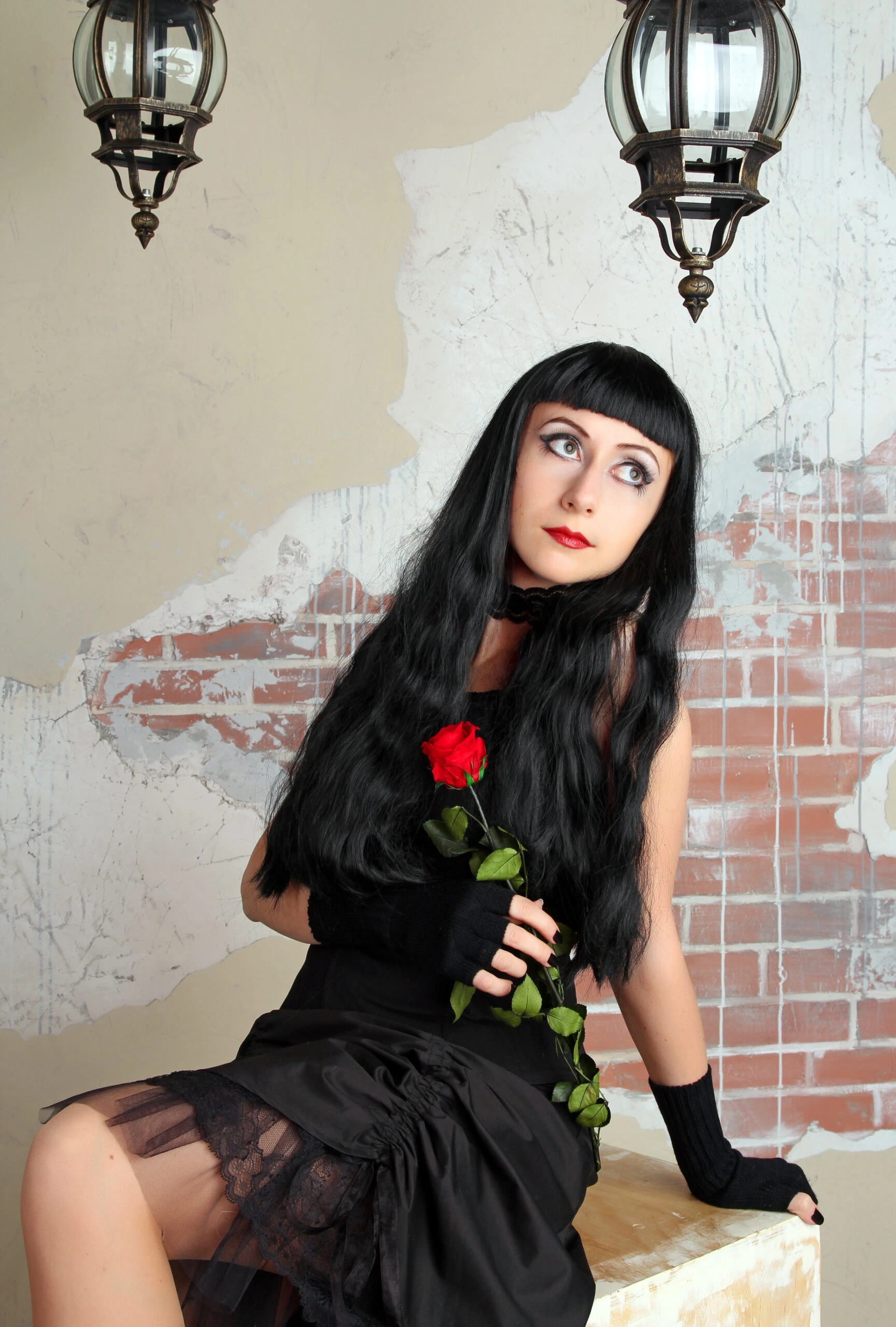 Gothic woman dressed in black, holding a red rose, with long black hair and dramatic makeup, sitting in front of a distressed wall.