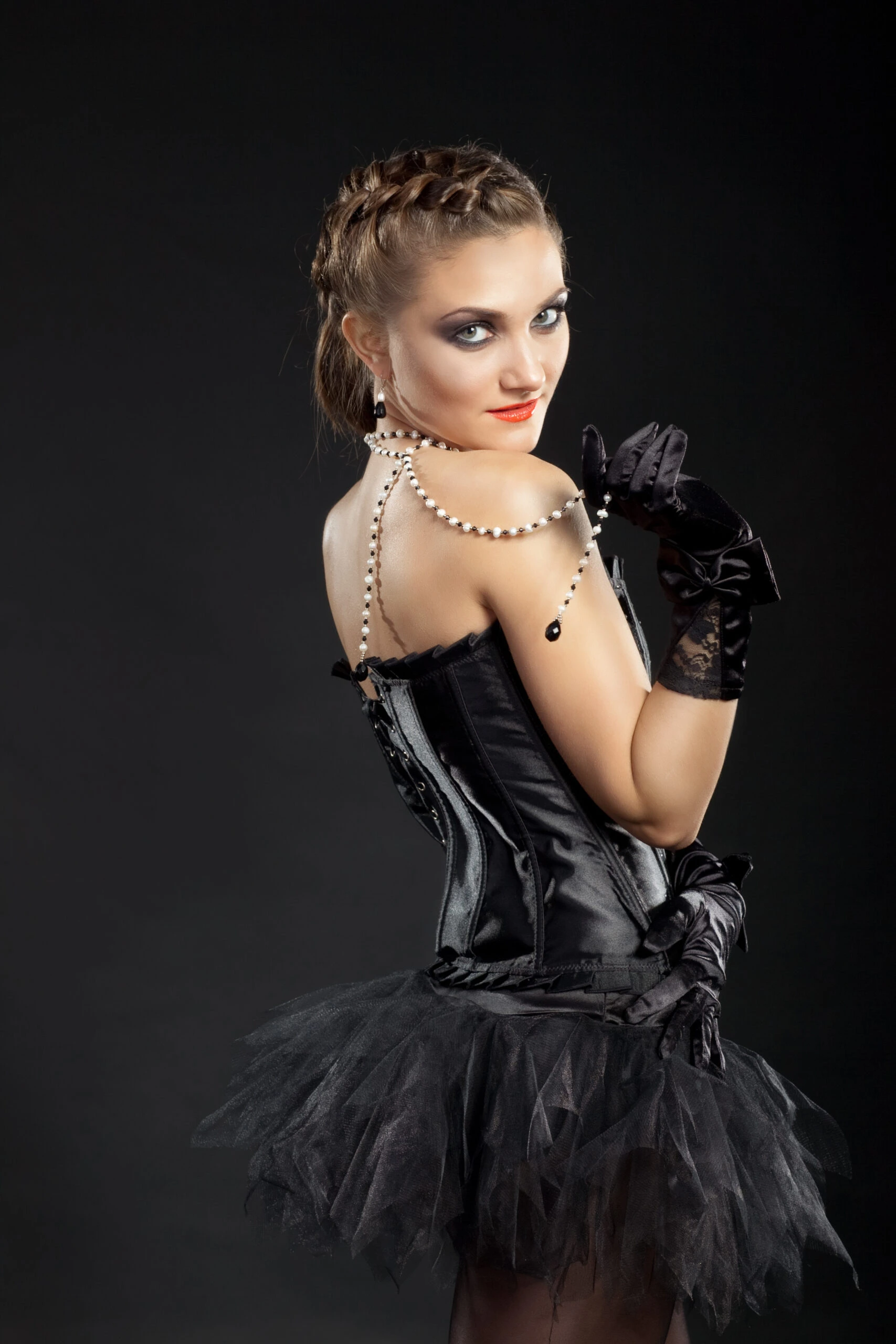 Woman in a black corset and tutu, wearing a pearl necklace, posing elegantly on a dark background.