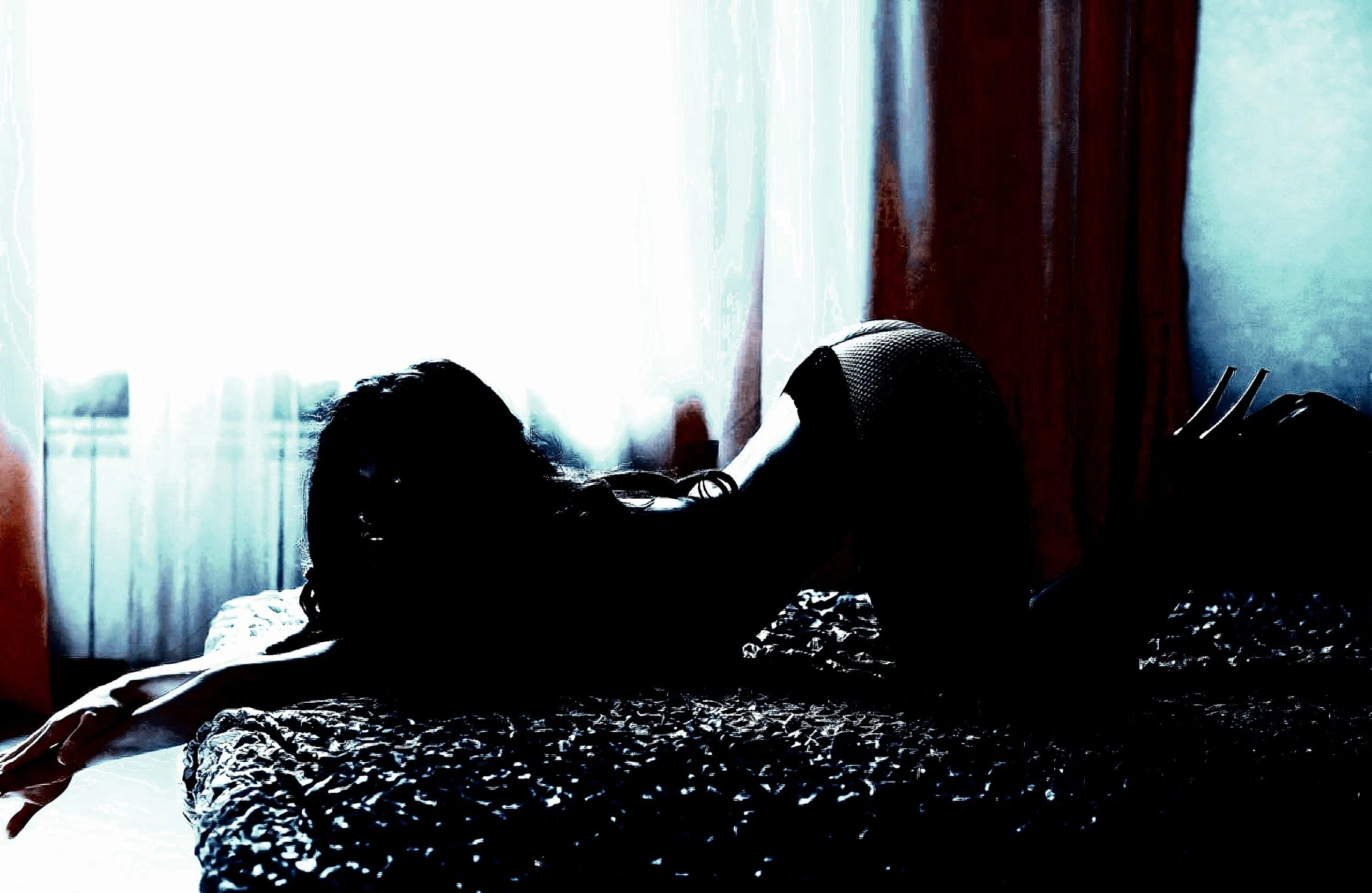 A woman in a seductive pose on a bed, lit by the soft glow of daylight coming through sheer curtains.